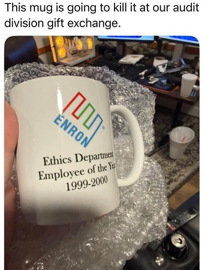 The image features a white ceramic mug held in someone's hand, partially wrapped in clear bubble wrap. The mug displays the Enron logo, a stylized depiction of three interconnected boxes in red, green, and blue. Below the logo, black text reads "Ethics Department Employee of the Year 1999-2000". The mug is positioned in the foreground, occupying the lower left portion of the frame.

The background is a blurred indoor scene, likely an office or home workspace. Visible elements include a desk wit…