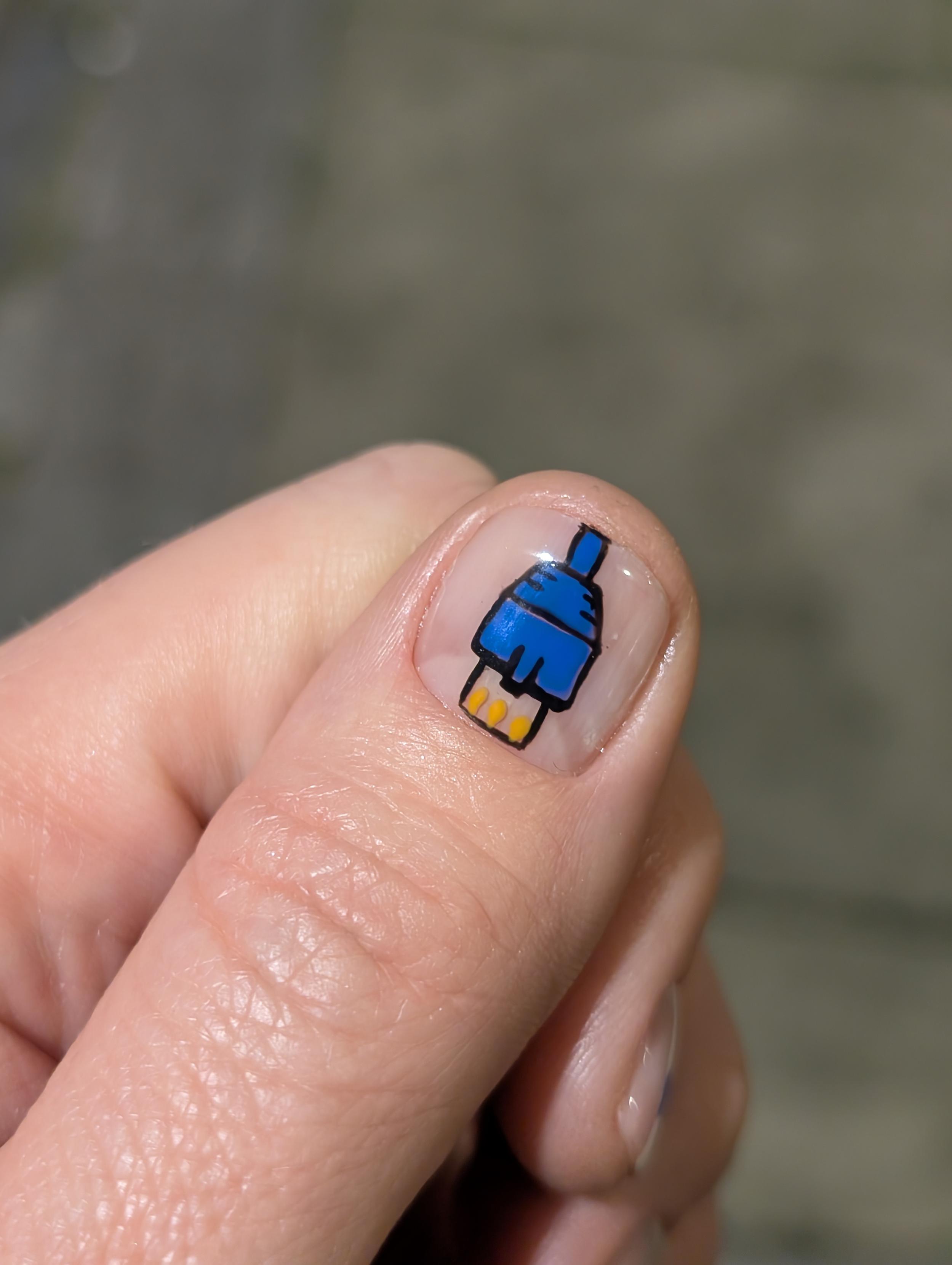 A close-up photo of a person's thumb featuring nail art of a blue RJ45 Ethernet plug. The design is hand-drawn with black outlines and three small yellow dots representing the connector pins at the tip, painted onto a clear nail base.