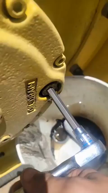 Video of someone draining the oil from a large machine. While they do so the drainage screw makes a sound while blowing bubbles. The mechanic stops then starts again which make the drainage hole make a screaming sound.