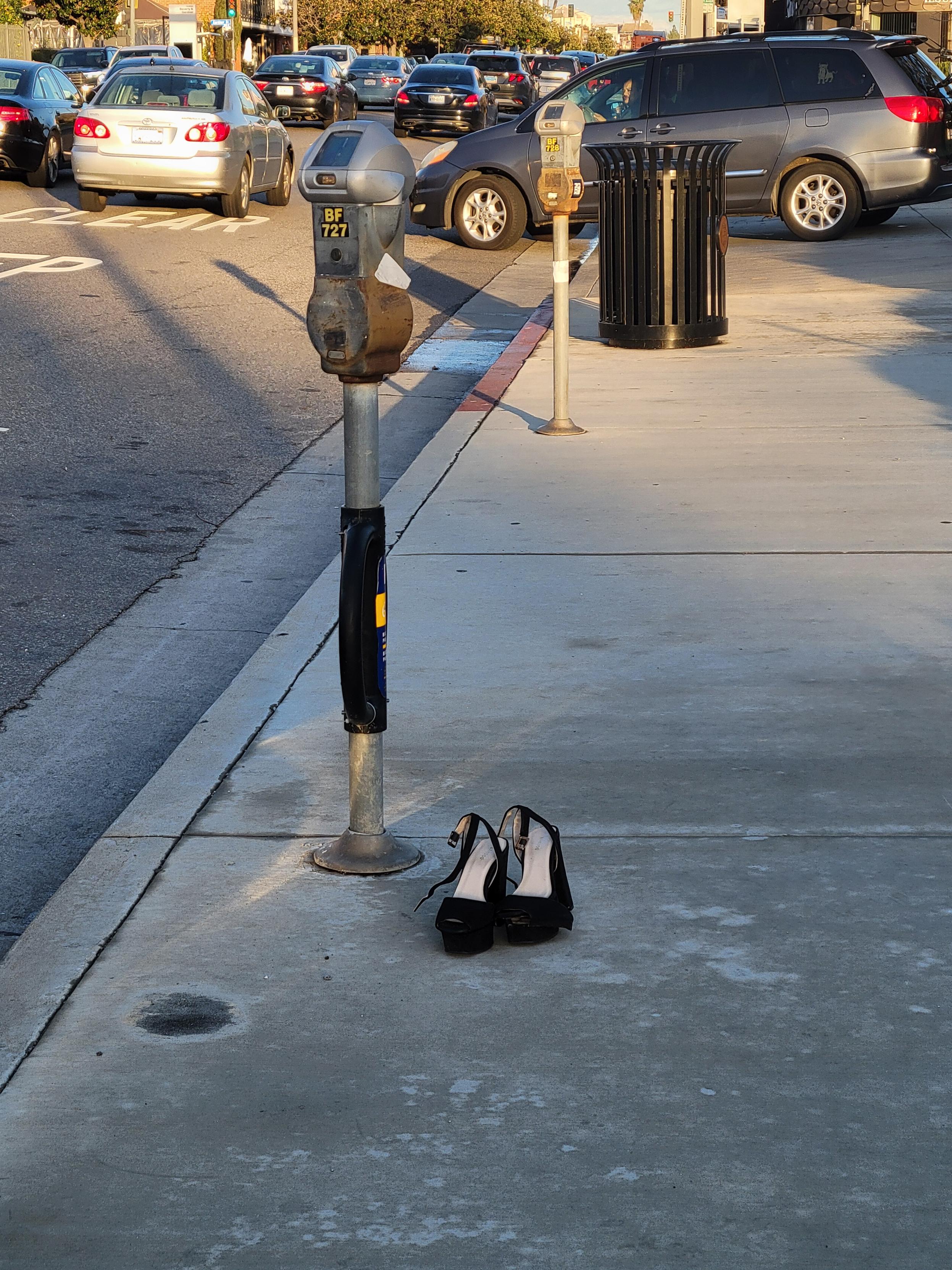 Urban sidewalk scene with a street filled with cars. On the sidewalk, next to a parking meter with a black pad wrapped around the pole, stands a pair of strappy black heels, unbuckled and left for some braver soul.