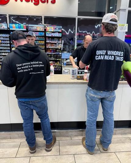 The image depicts a scene inside a convenience store, likely a gas station or a similar establishment, with a counter area prominently featured. Three men are visible, two of whom are customers standing at the counter, and one is an employee behind it. The perspective is from behind the two customers.

The customer on the left wears a black hooded sweatshirt and blue jeans. The back of his sweatshirt has white text printed on it, reading "Dear person behind me, the world is a better place with …