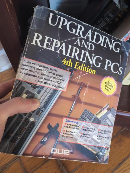 A hand holds a worn book titled "UPGRADING AND REPAIRING PCS 4th Edition" by QUE. The cover features large white text for the title with "4th Edition" in yellow, a red-bordered quote from Info World Magazine stating "...an exceptional book, covering most of what you'd ever need to know about how to upgrade and repair a PC.", and a yellow circular badge that reads "Over 650,000 Copies Sold!". Below the title, a red-bordered section lists "Learn to:" with bullet points: "Add Memory and Disk Drive…