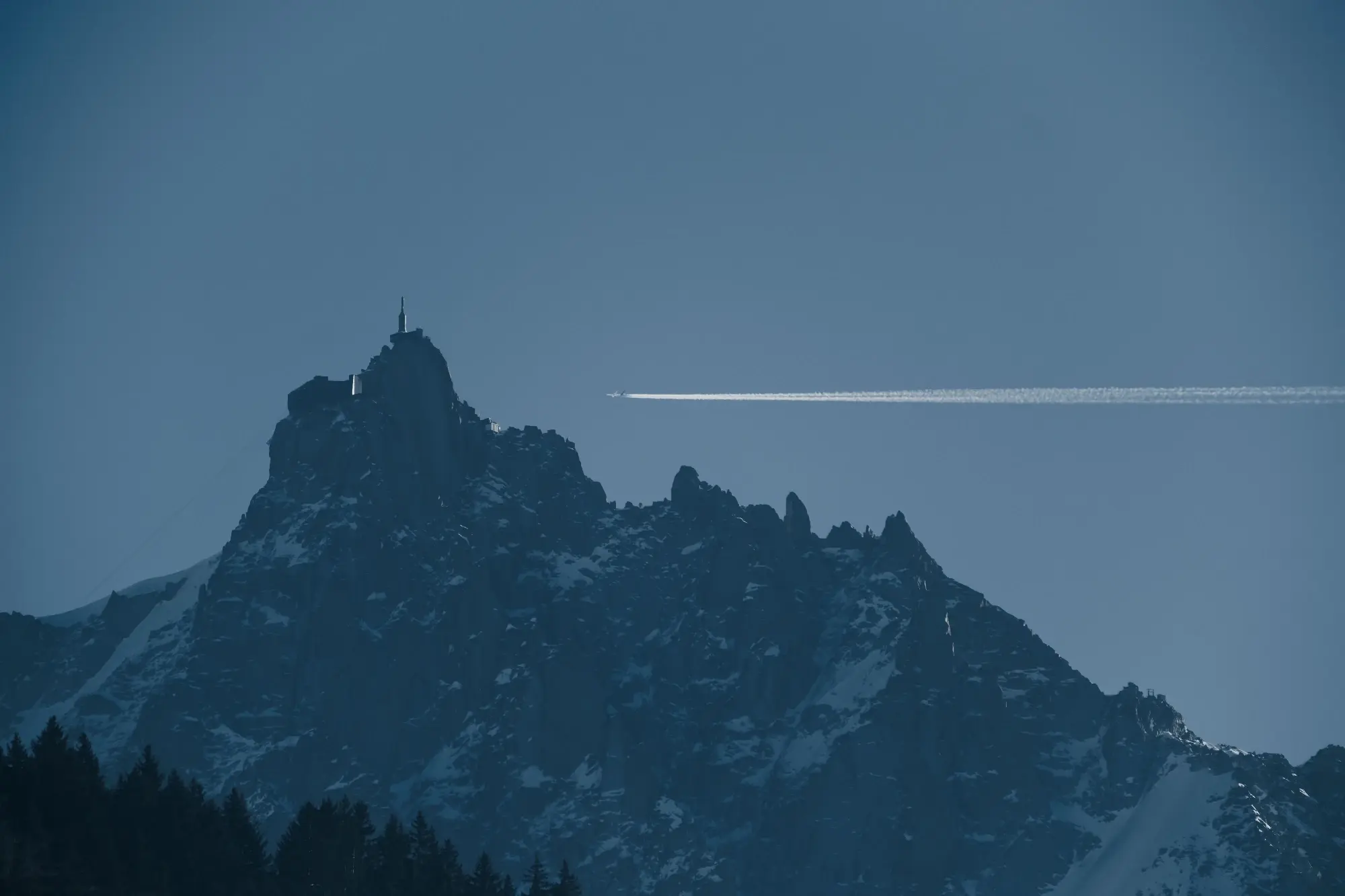 Photo of a mountain top with a building at the peak.
In the background there is a vast empty sky and a plane flying from the right side of the frame leaving a horizontal trail towards the mountain top.