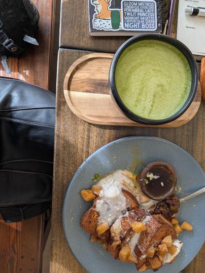 Top down photo of a plate of french toast, a large cup of matcha, and a notebook on a table. The lap of a black leather skirt is visible at the edge of the table