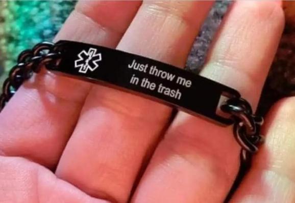 The image is a close-up photograph focusing on a black medical alert bracelet worn on a person's hand. The bracelet consists of a black metal chain and a central black rectangular bar. On the bar, there is a white engraving of the Star of Life, a widely recognized medical symbol, positioned on the left side. To the right of the symbol, the phrase "Just throw me in the trash" is engraved in a clean, white, sans-serif font, arranged in three lines. The bracelet is wrapped around the person's wris…