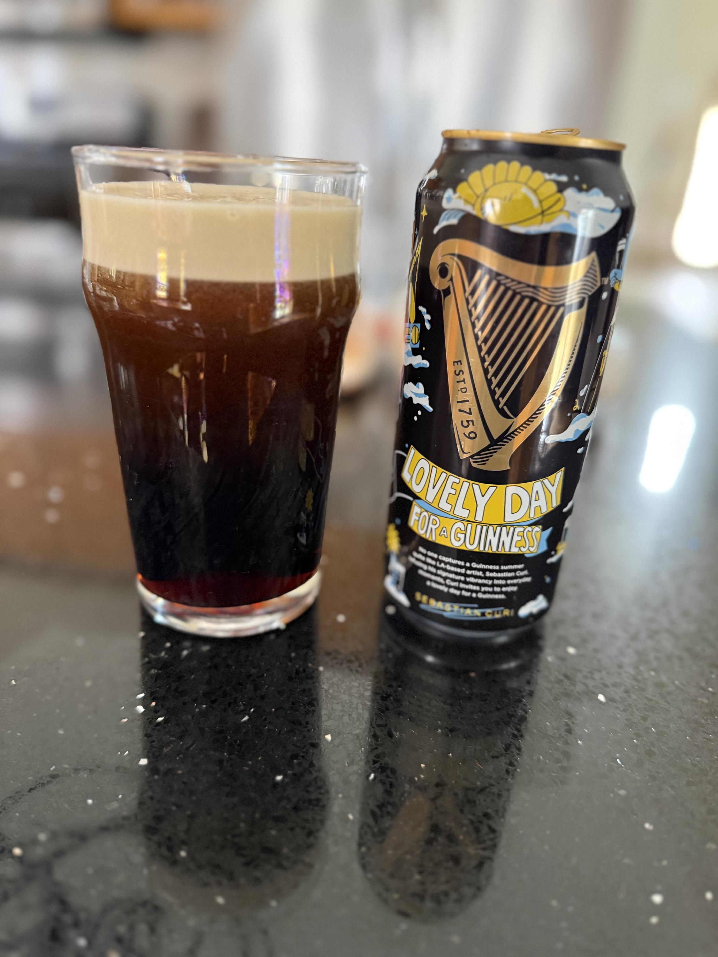 A pint glass full of dark beer with a tan head. The black pint can has a gold harp and the words Lovely Day for a Guinness in white letters on a yellow banner 