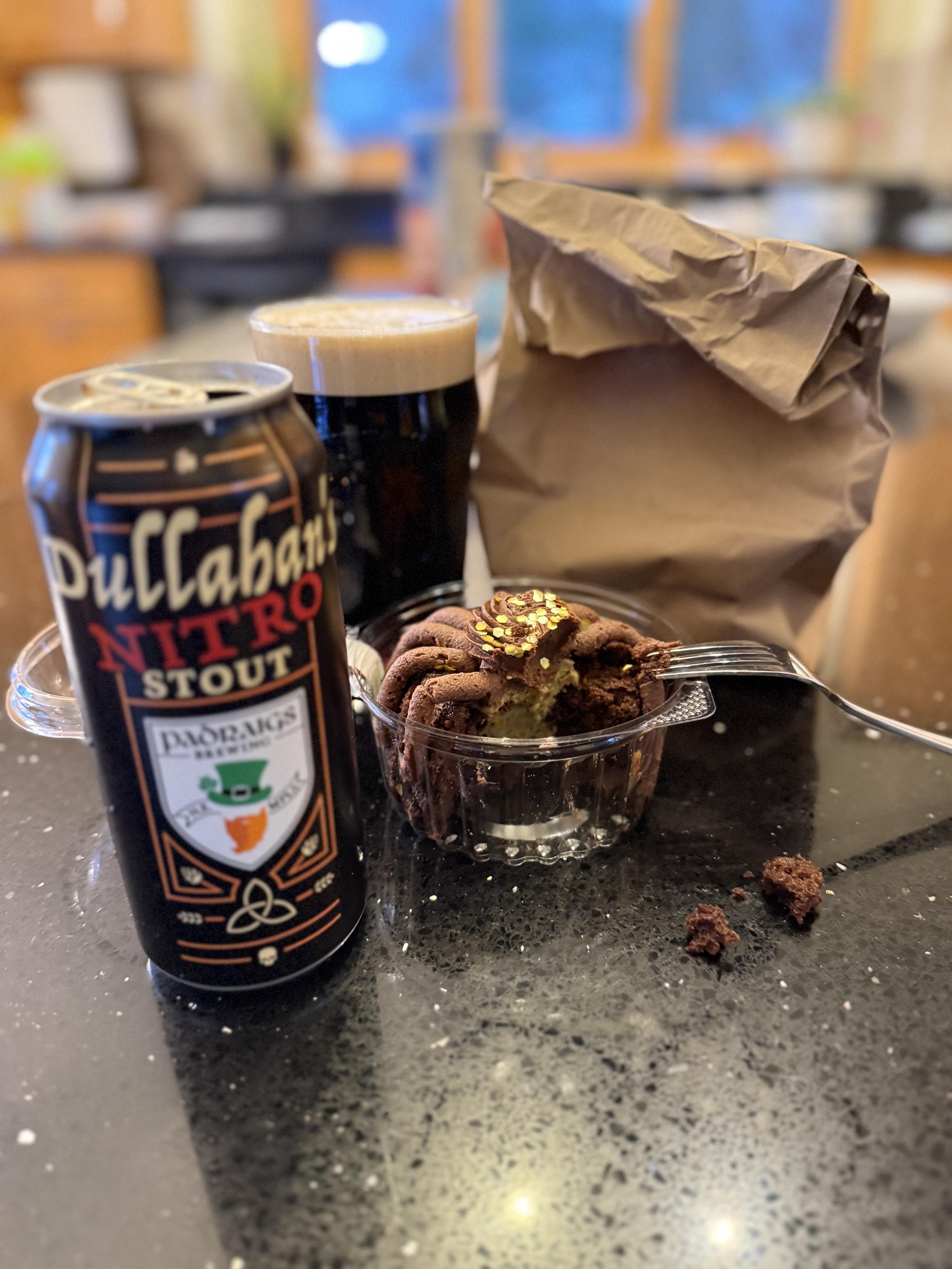 A brown pint can is next to a full pint glass of dark beer with a tan head. A clear container holds a light brown cake with a bite taken.  A brown paper bag conceals (perhaps?) another cake.