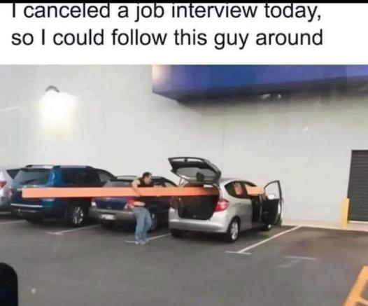 The image is a meme overlayed with text that reads "I canceled a job interview today, so I could follow this guy around." Below the text is a photograph taken in what appears to be a parking lot, likely outside a large store or warehouse.

In the foreground, a person is struggling to load a very long, bright orange plank of wood into the back of a silver car. The car's trunk is open, and the plank extends all the way through the interior, with its front end protruding out of the open driver's s…