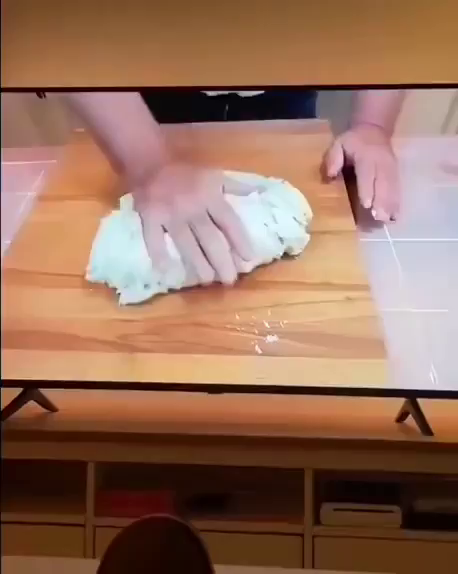 Video showing a cooking TV show with someone kneading bread dough, then the camera pans over to a cat making biscuits on a blanket while watching that cooking show.