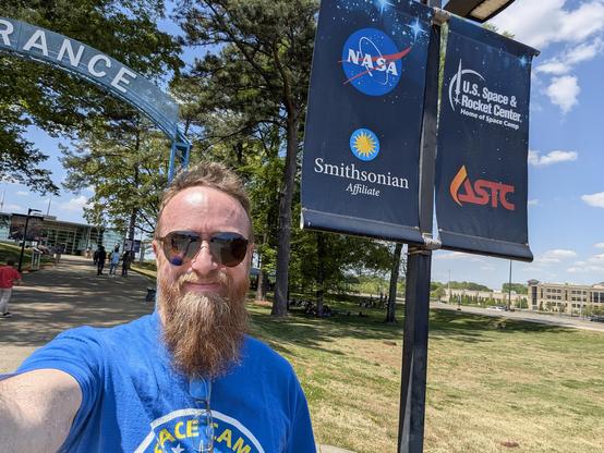 A person with a beard and sunglasses takes a selfie outdoors in front of a signpost on a sunny day. They are wearing a blue shirt with a partially visible yellow logo. Behind them to the right, two vertical blue banners hang from a pole. The left banner displays the NASA logo and the text "Smithsonian Affiliate". The banner next to it displays the text "U.S. Space & Rocket Center", "Home of Space Camp", and the orange "ASTC" logo. In the background, a large metal archway shows the letters "RANC…