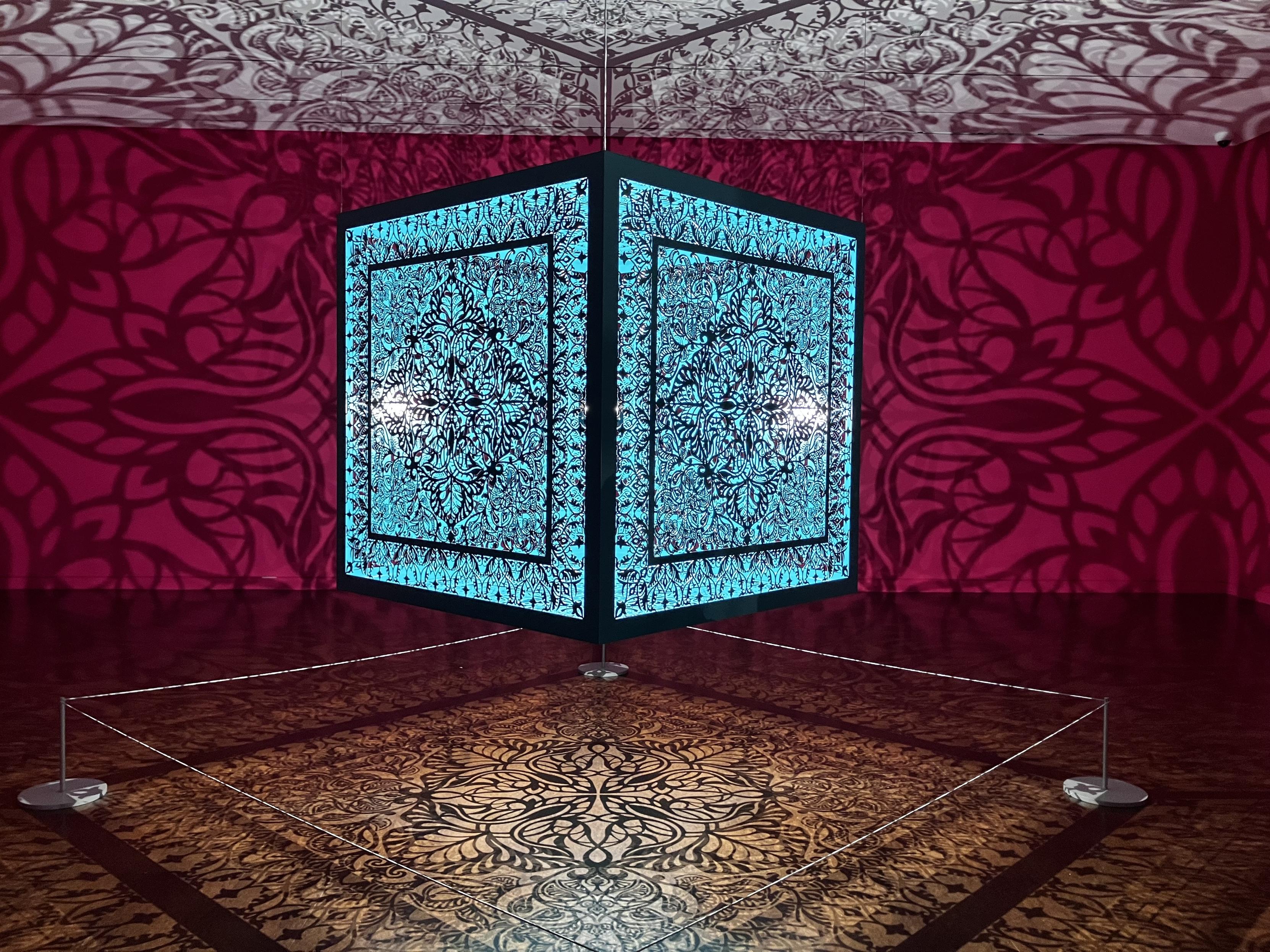 In the center of the image is a blue cube, illuminated from inside. The cube faces are intricately patterned, and the patterns are projected below, on a white/yellow surface; above, on a pure white surface; and on the surrounding walls, which are a rich red.