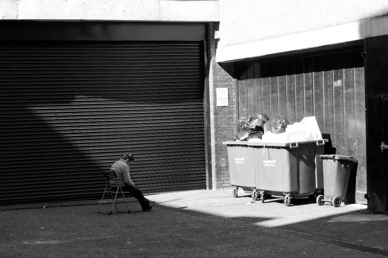 A man sitting in shadow next to a massive garage gate in front of sunlit trash bins.