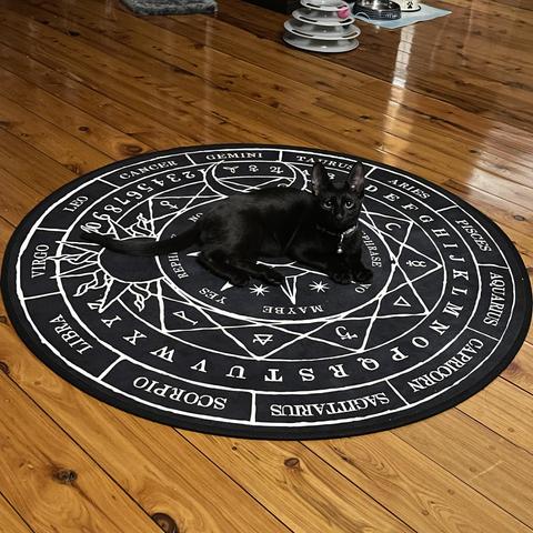 Photo of a young black cat laying down on a Ouija board type, circular rug. The rug has zodiac signs, letters, numbers, sun & moon symbols, etc.
