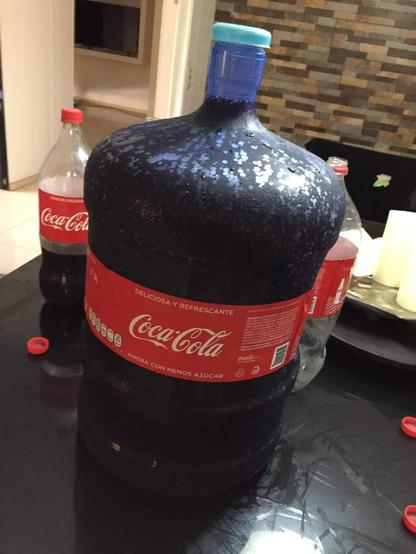 Photo of a 5 gallon (~19 liters) office water jug filled completely with Coca-Cola, soda drink label included (in Spanish). On the table near by rests an empty 2 liter and a mostly empty 2 liter bottles of Coke. Wonder how many calories that contains?