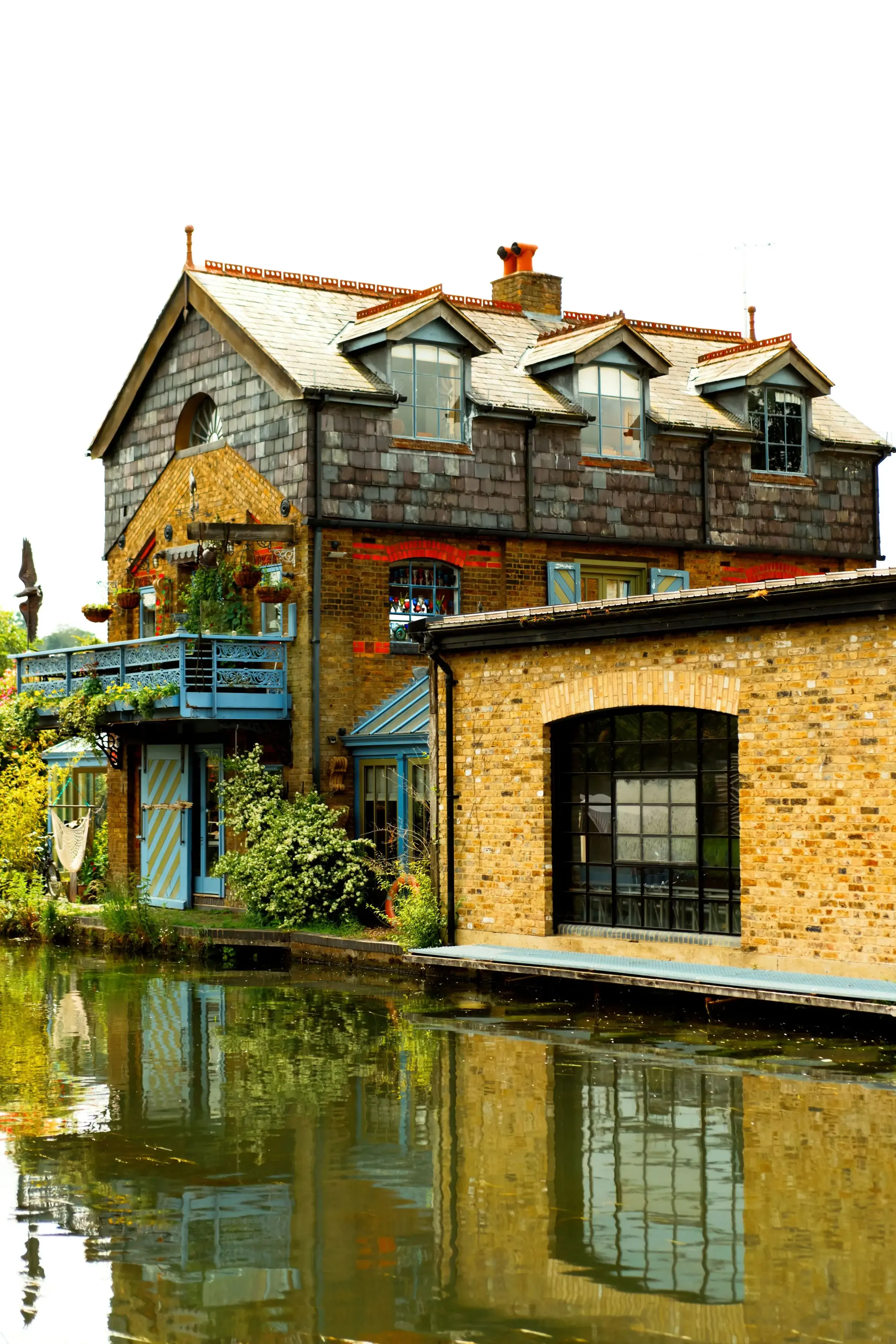 A multi-story brick house with slate shingles and dormer windows sits along the Grand Union Canal in Berkhamsted, Hertfordshire. The building features a prominent blue wooden balcony adorned with hanging plants, located above light blue doors with diagonal yellow stripes. To its right, a lower yellow brick building with a large arched window is situated at the water's edge. The still water of the canal reflects the detailed architecture and surrounding greenery under a bright sky.