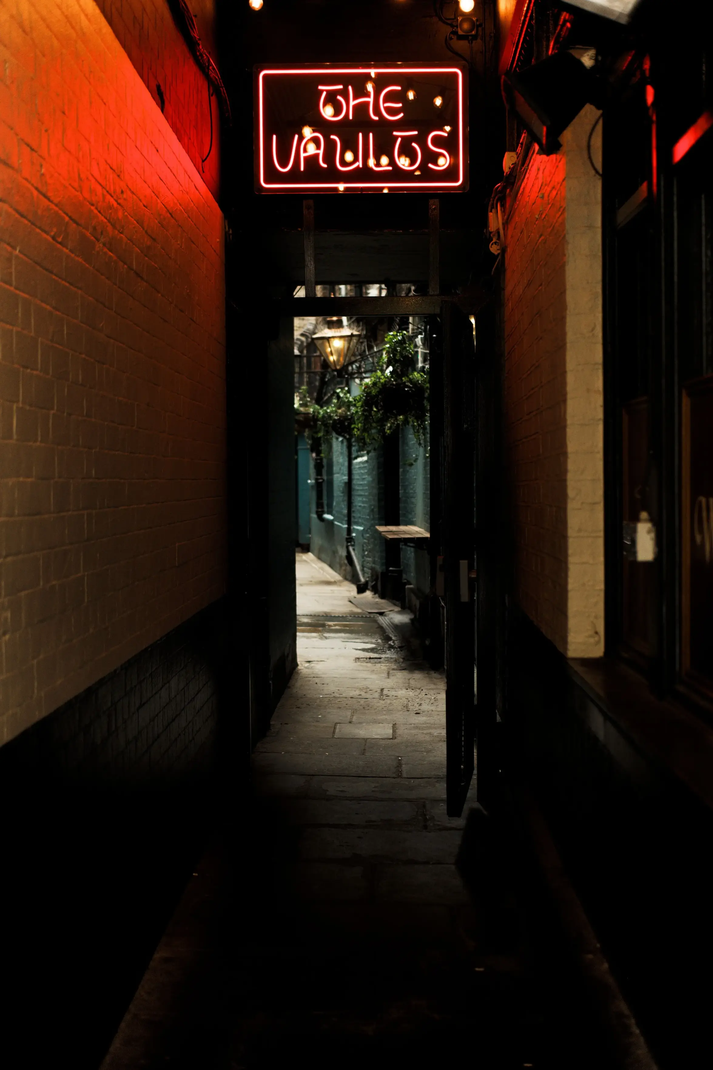 A dark, narrow alleyway at night featuring brick walls on both sides. A glowing red neon sign with stylized lettering reading "THE VAULTS" hangs above an open doorway, casting a vibrant red light down the passageway.