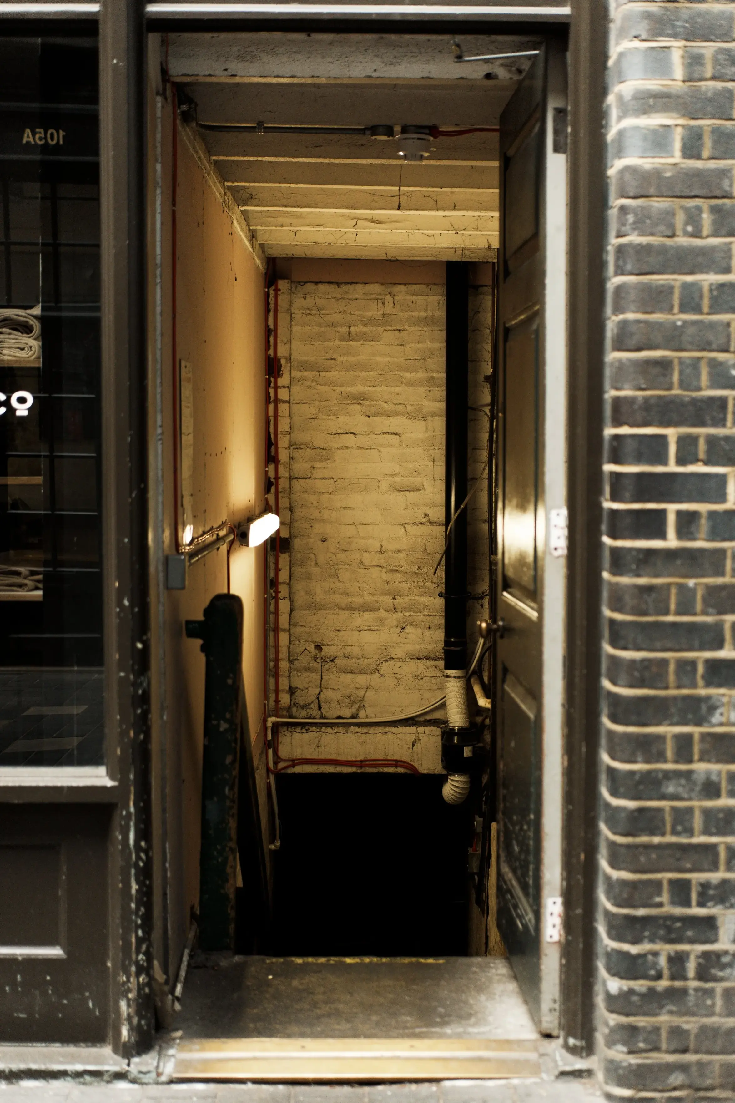 You said: write alt textAn open doorway leads into a dimly lit, narrow indoor passageway. The doorway is framed by a dark glass-paneled storefront on the left and a weathered, exposed brick wall on the right. Inside, the corridor features light-colored walls illuminated by warm light fixtures, a white-painted brick accent wall at the back, a vertical pipe, and a short flight of stairs leading downward.