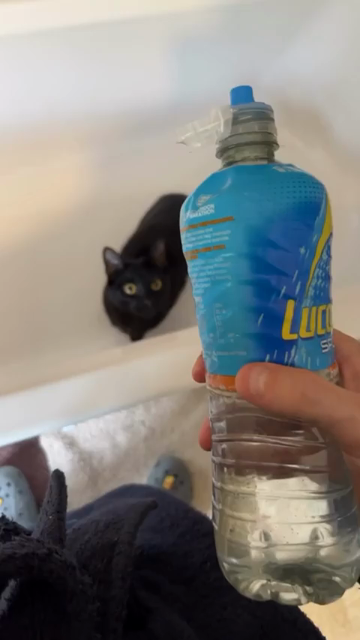 Video of someone wearing crocs and holding a sports water bottle looking down at a black cat in the tub who is staring at the bottle. The human slowly starts to point the bottle downward while the kitty's eyes dilate with excitement. They then squirt the bottle around the cat as the cat happily tries to catch the water. It's a silly game this cat seems to absolutely love for sure!