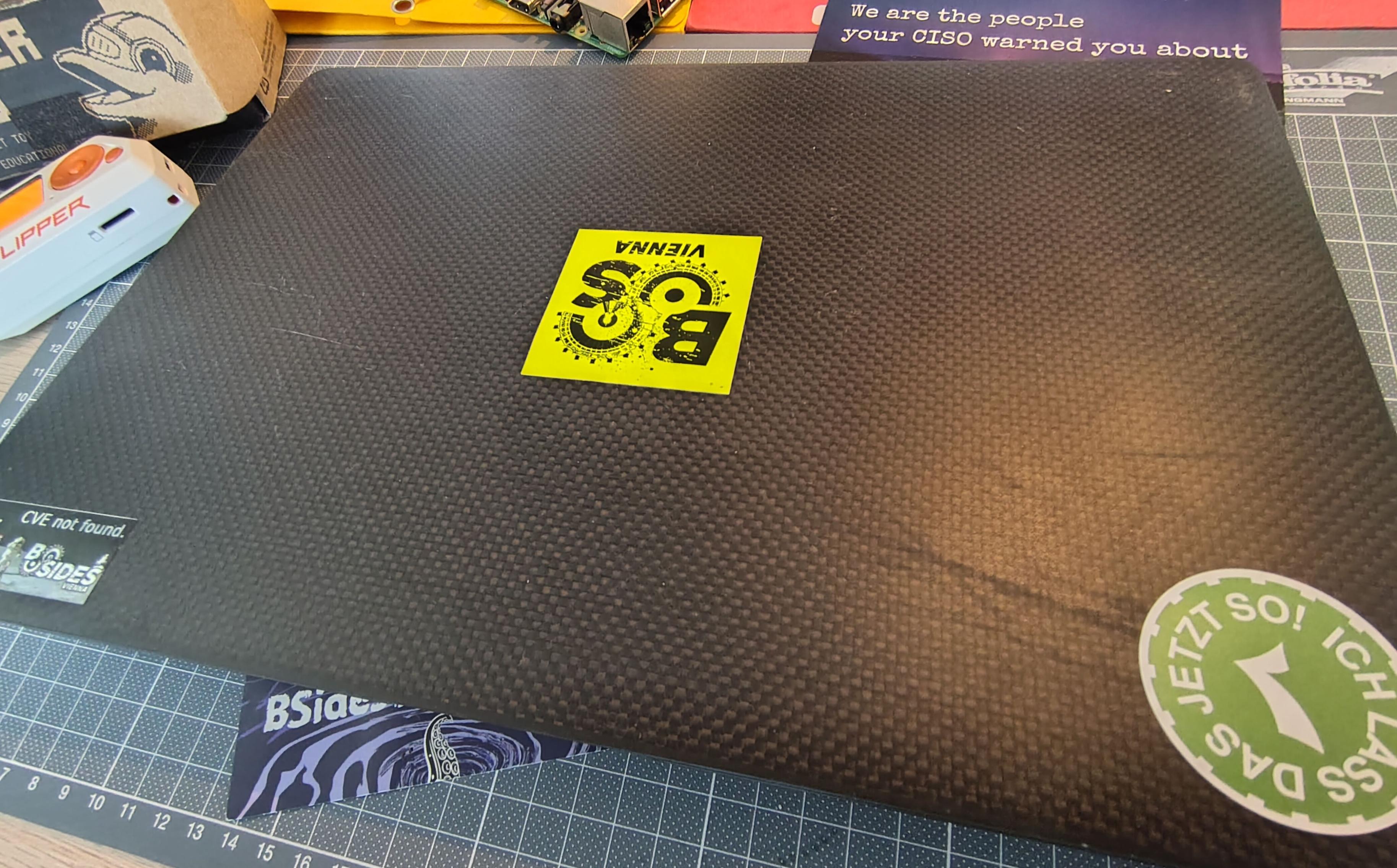Picture of a Laptop with two BSidesVienna stickers and another sticker which says "Ich lass das jetzt so". A flipper is visible on the left, a raspberry on top and a card saying: "We are the people your CISO warned you about." on the top right.