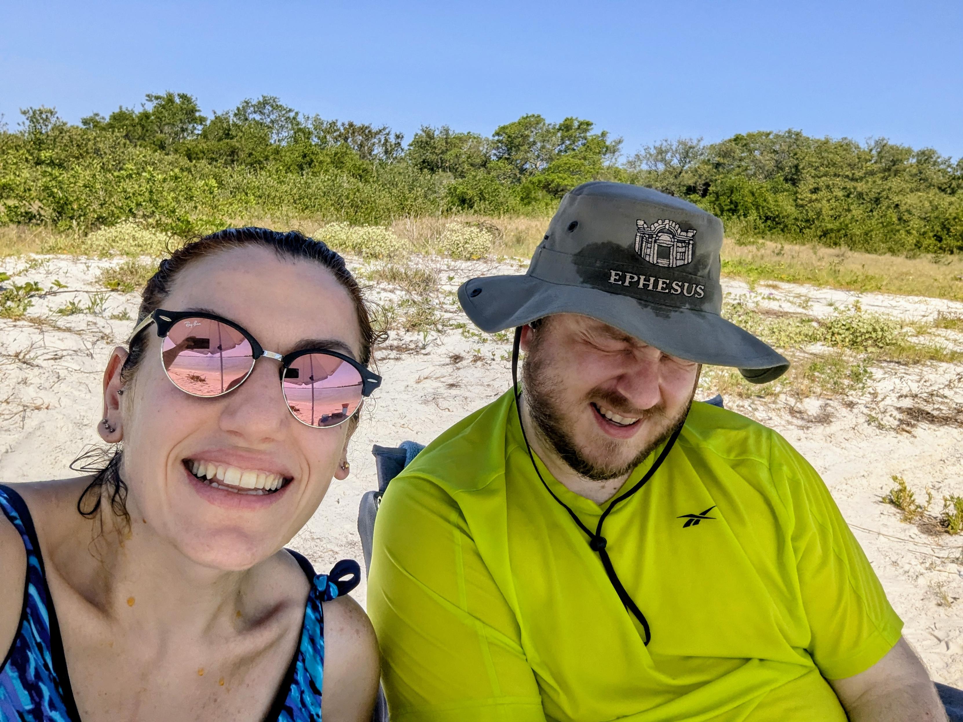 Two people at the beach. My wife is happy wearing sunglasses, while I have my eyes squashed shut with a painful wince on my face.