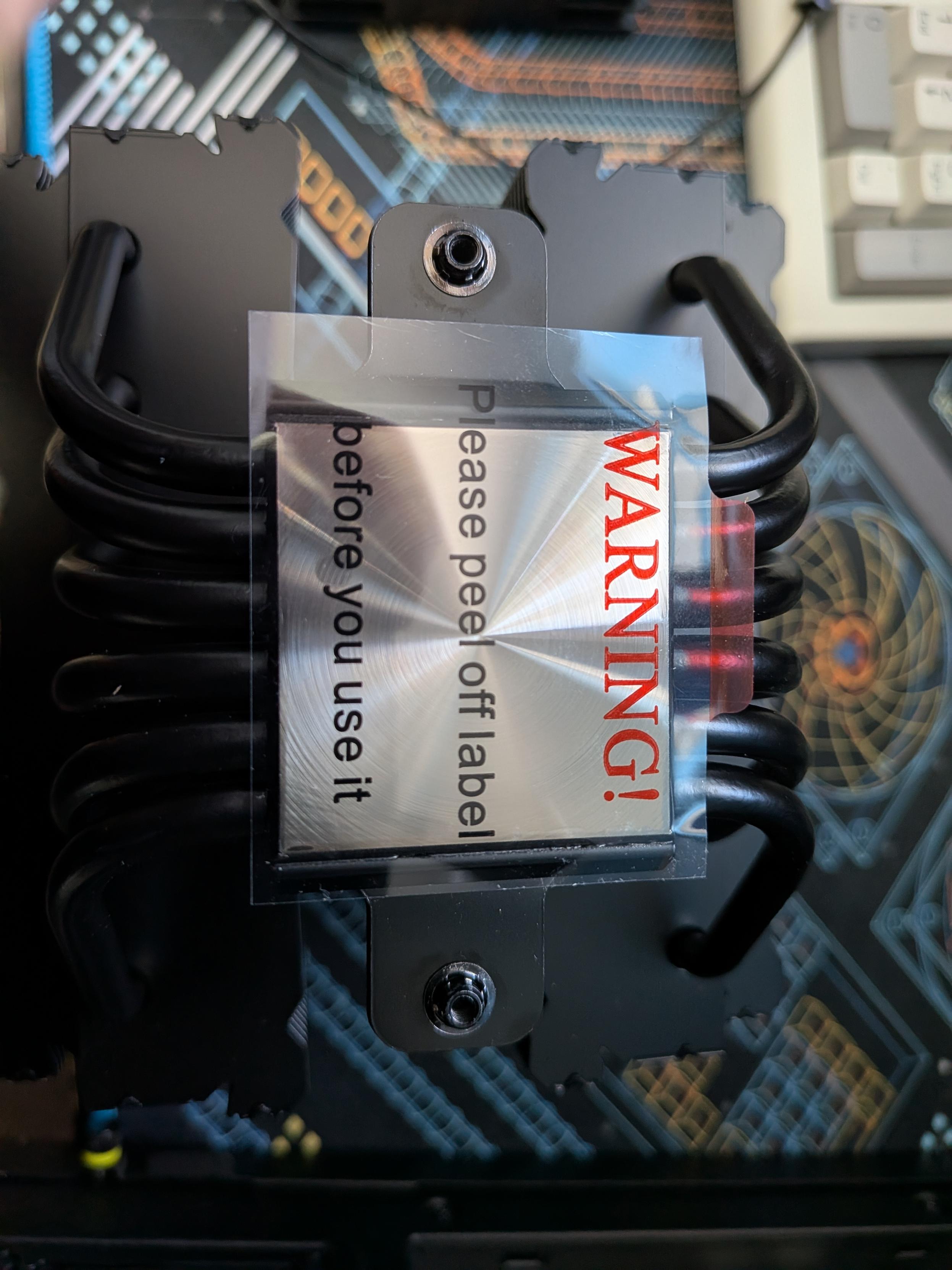 An overhead close-up view of a black CPU air cooler mounted on a motherboard. A clear plastic protective film covers the flat metal base of the cooler. The text on the film reads, "WARNING! Please peel off label before you use it". The cooler features several thick, curved black heat pipes. Beneath the cooler is a motherboard with blue and white circuit patterns, and a portion of a light-colored computer keyboard is visible in the top right corner.
