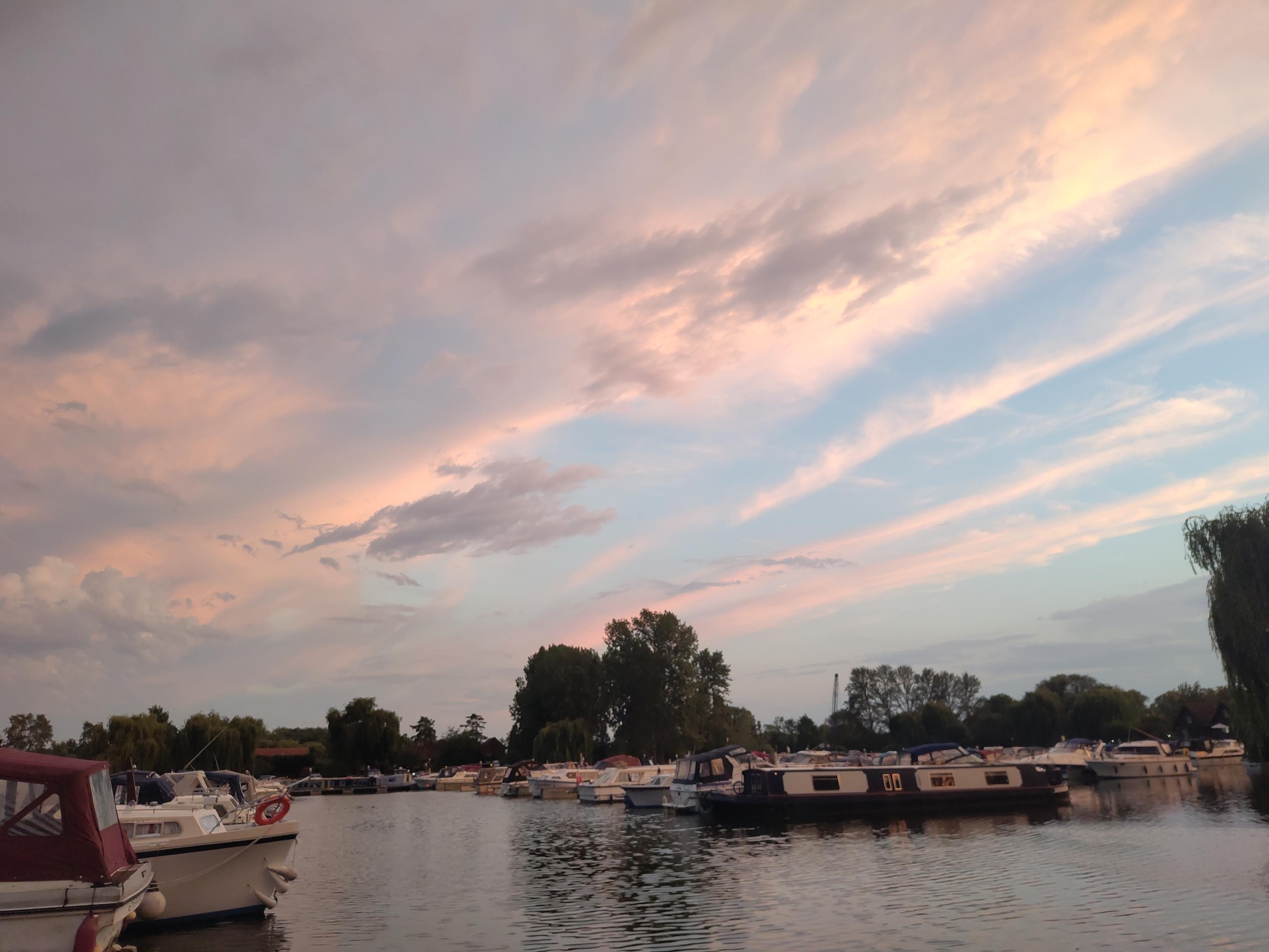 Azure blue skies with bands of high cloud shading to pink, above a marina with trees on the horizon and canal boats and cruisers in the foreground.
