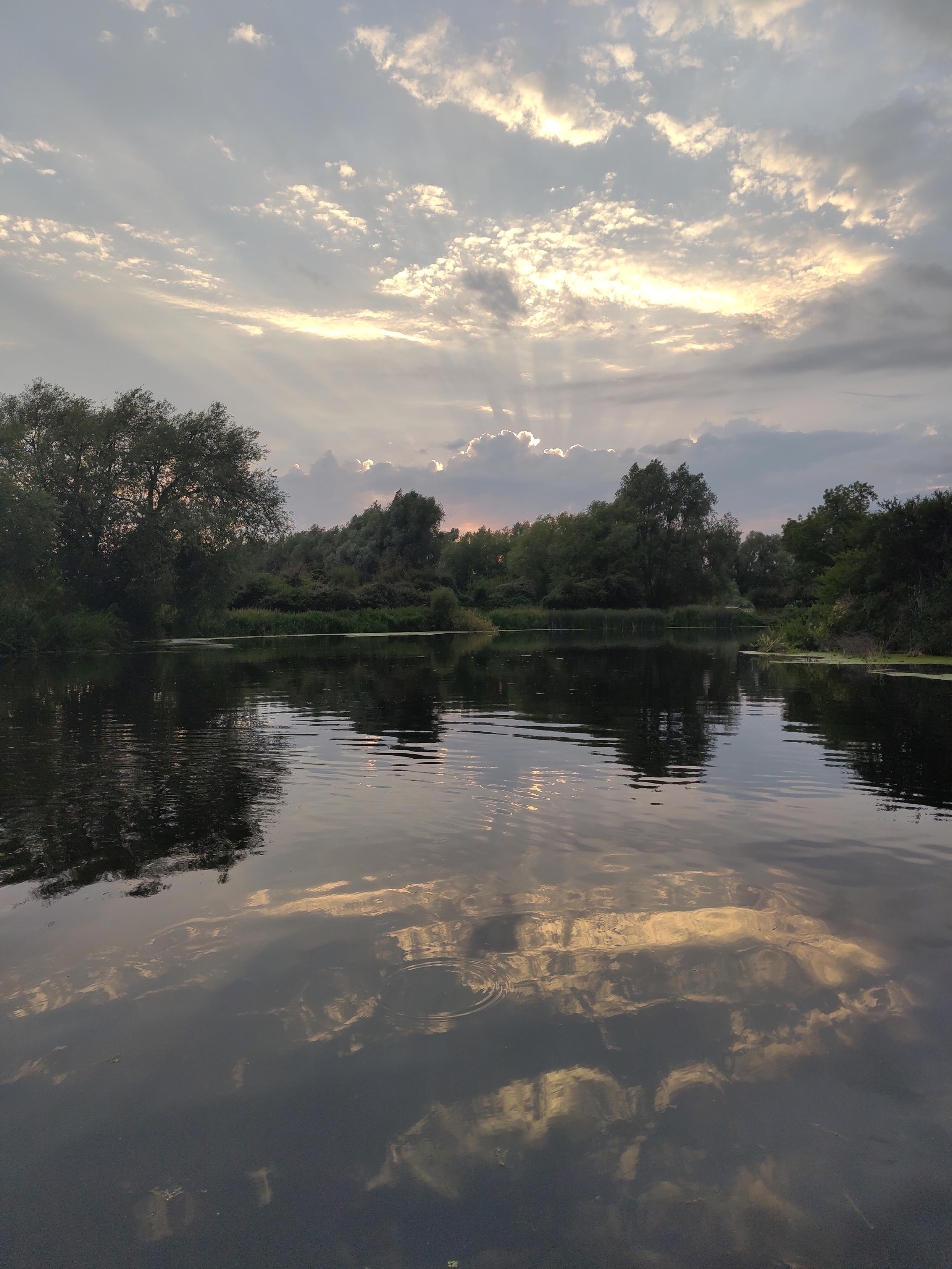 Evening sky over a river, with light steaming through high white clouds, reflected in the water of the river below.