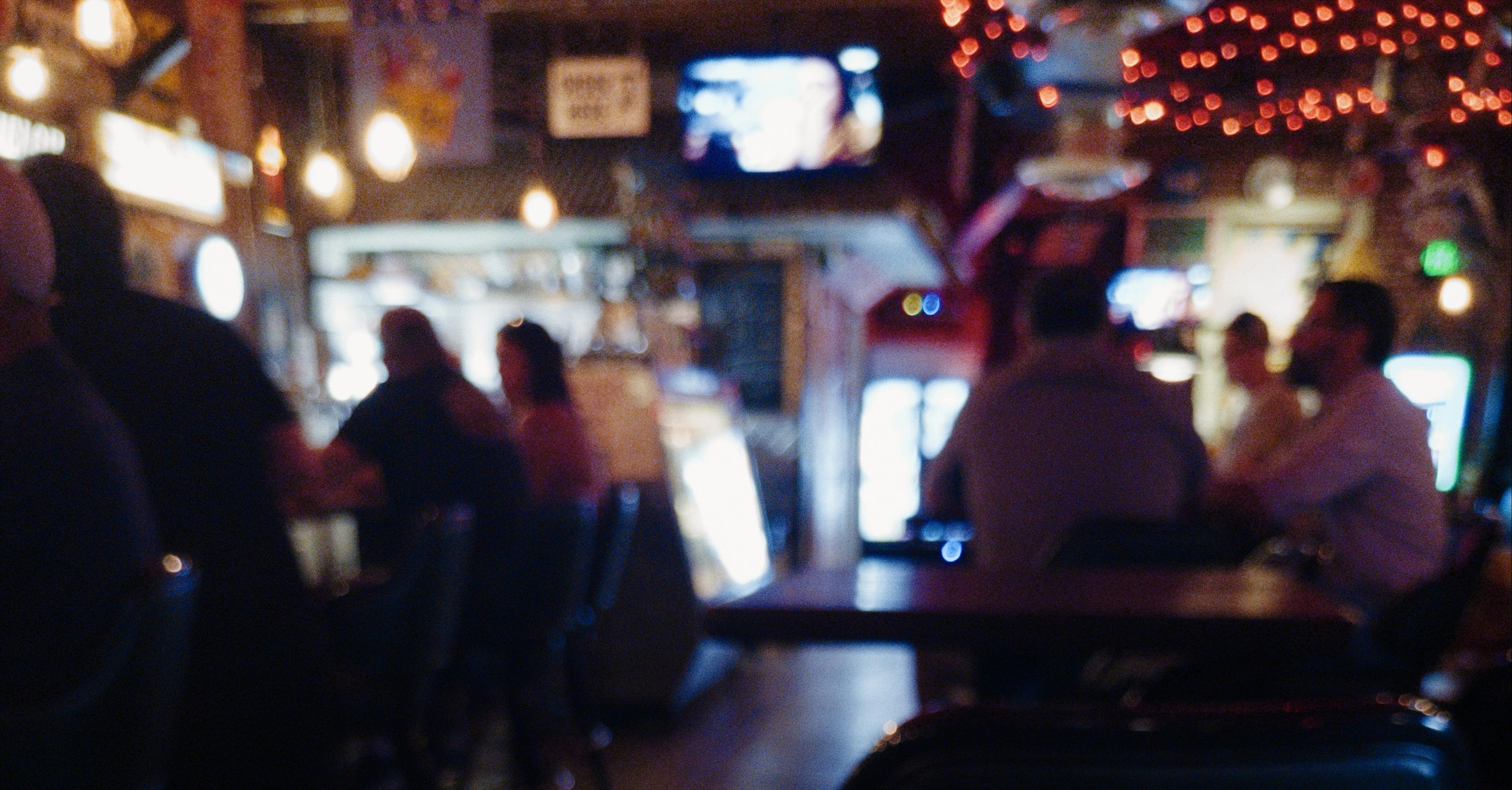 A dimly lit bar scene with blurred figures seated at a counter. Television and faint colorful lights are visible in the background.
