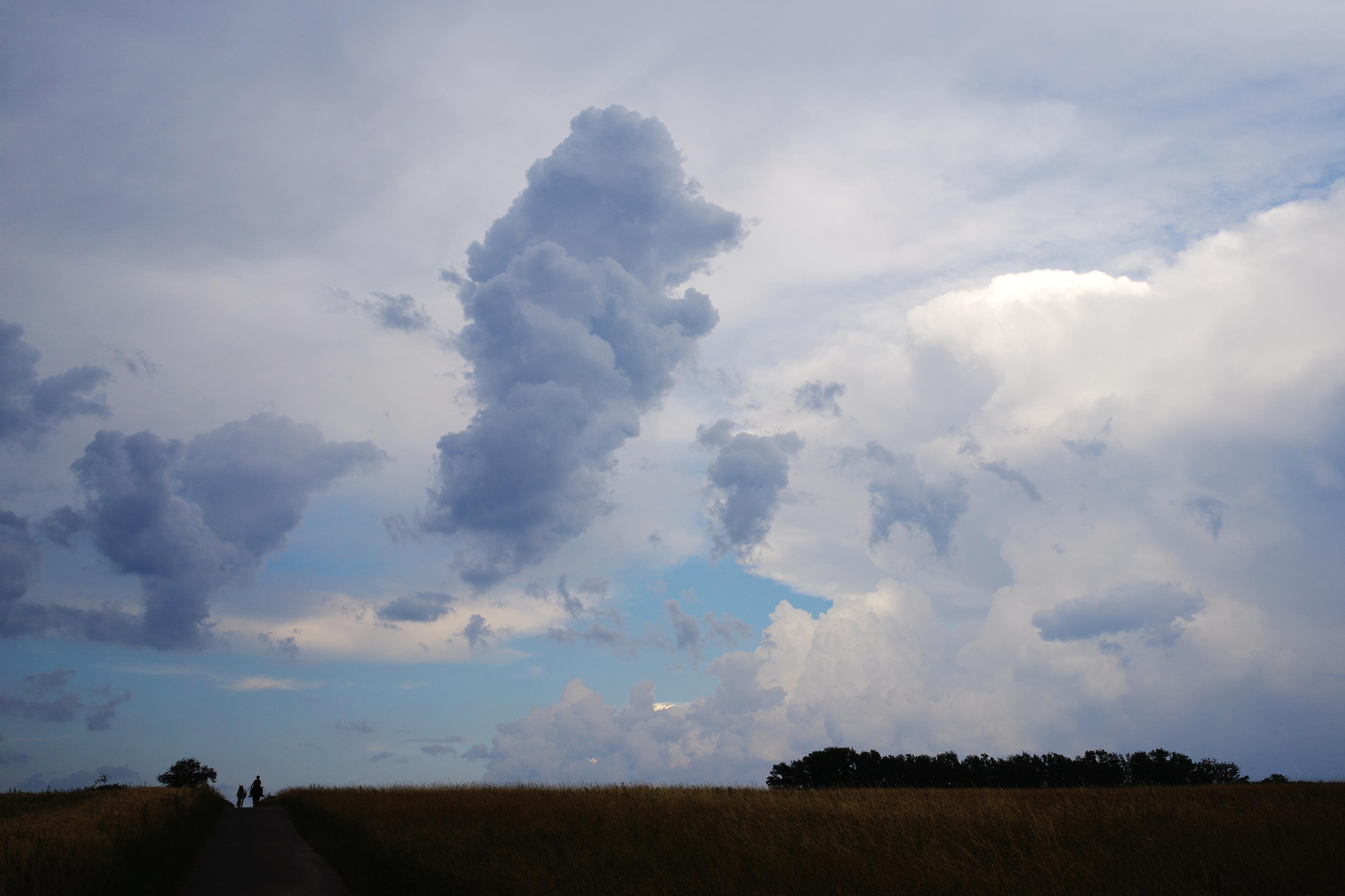 A cloudscape photo with a little bit of fields and trees at the bottom of the frame. In the lower left corner a small rider leading another horse can be seen.