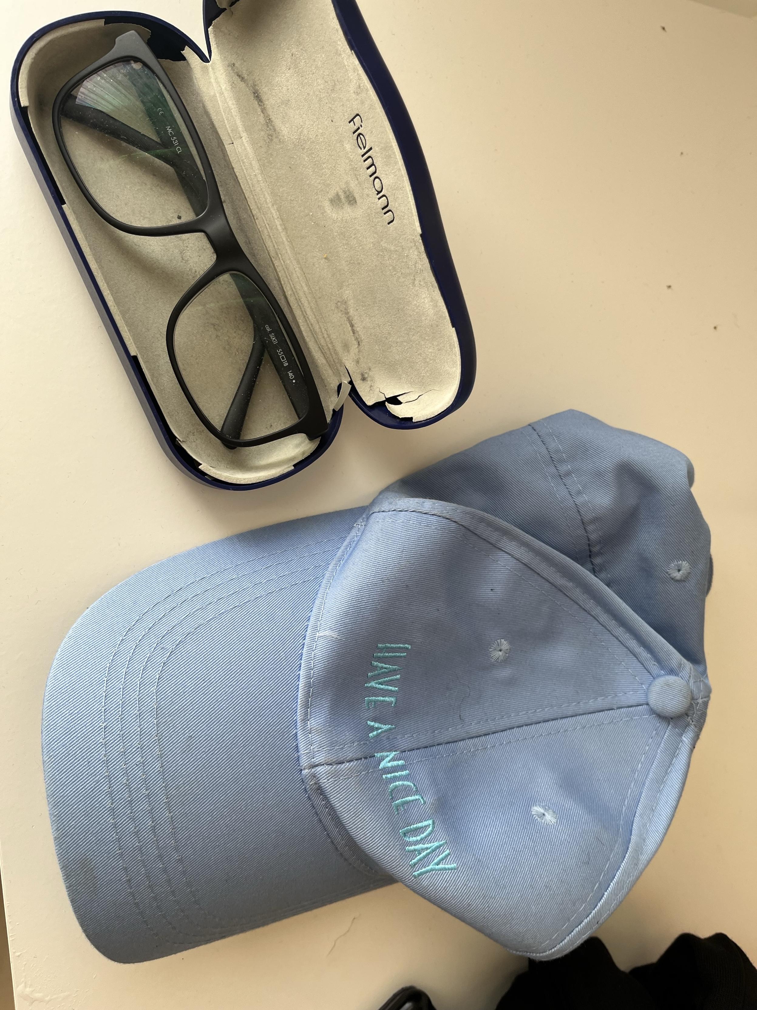 Blue baseball cap with text saying „have a nice day“, and a black pair of glasse in a case from Fielmann