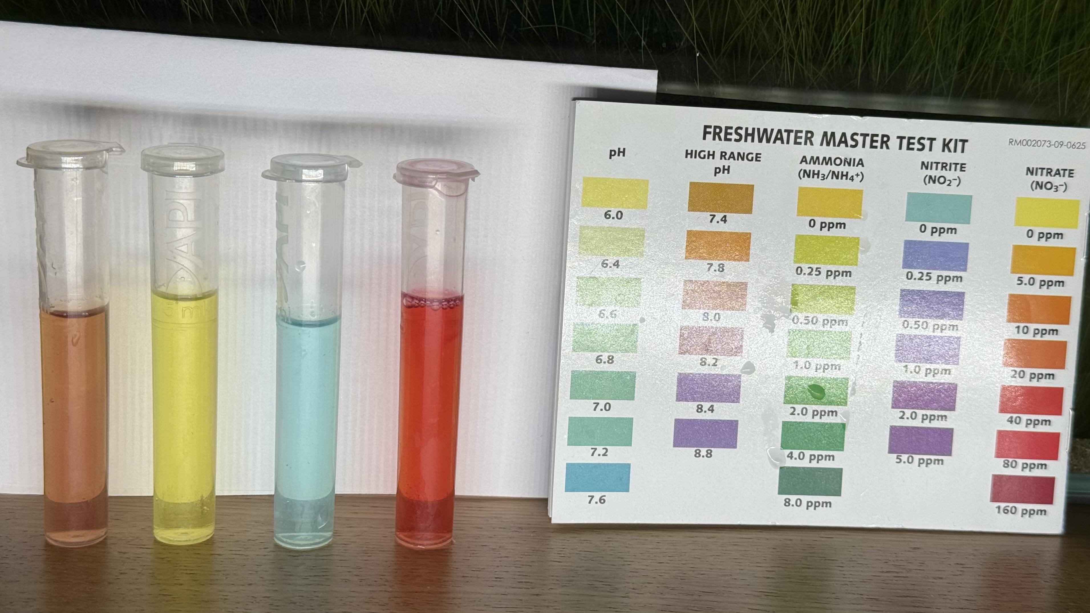 An API Freshwater Master Test Kit, showing four plastic test tubes lined up next to the color comparison chart. From left to right, the tubes read: an orange tube (pH, reading low on the scale), a bright yellow tube (ammonia at 0 ppm), a pale blue-green tube (nitrite at 0 ppm), and a red-orange tube (nitrate, reading roughly 20–40 ppm). The printed reference card on the right shows color gradients for pH, high range pH, ammonia, nitrite, and nitrate.