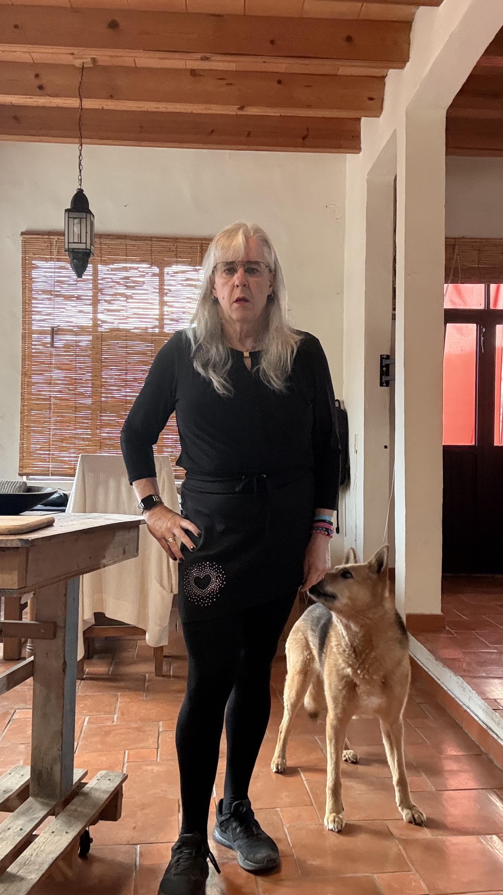 A woman with long gray hair and glasses stands indoors with a German Shepherd mix beside her. She is wearing black tights, a small black skirt, and a black top with three-quarter sleeves.