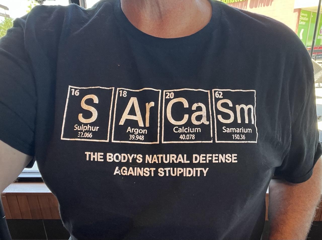 Selfie style pic of the top part of a black t-shirt with white lettering worn by a white male that shows elements (from the table of elements) lined up so that it spells.  Sarcasm. Below that is reads THE BODY'S NATURAL DEFENSE AGAINST STUPIDITY.  

The elements boxes include the following:

16 S Sulphur 32.066 

18 Ar Argon 39.948 

20 Ca Calcium 40.078 

62 Sm Samarium 
150.36