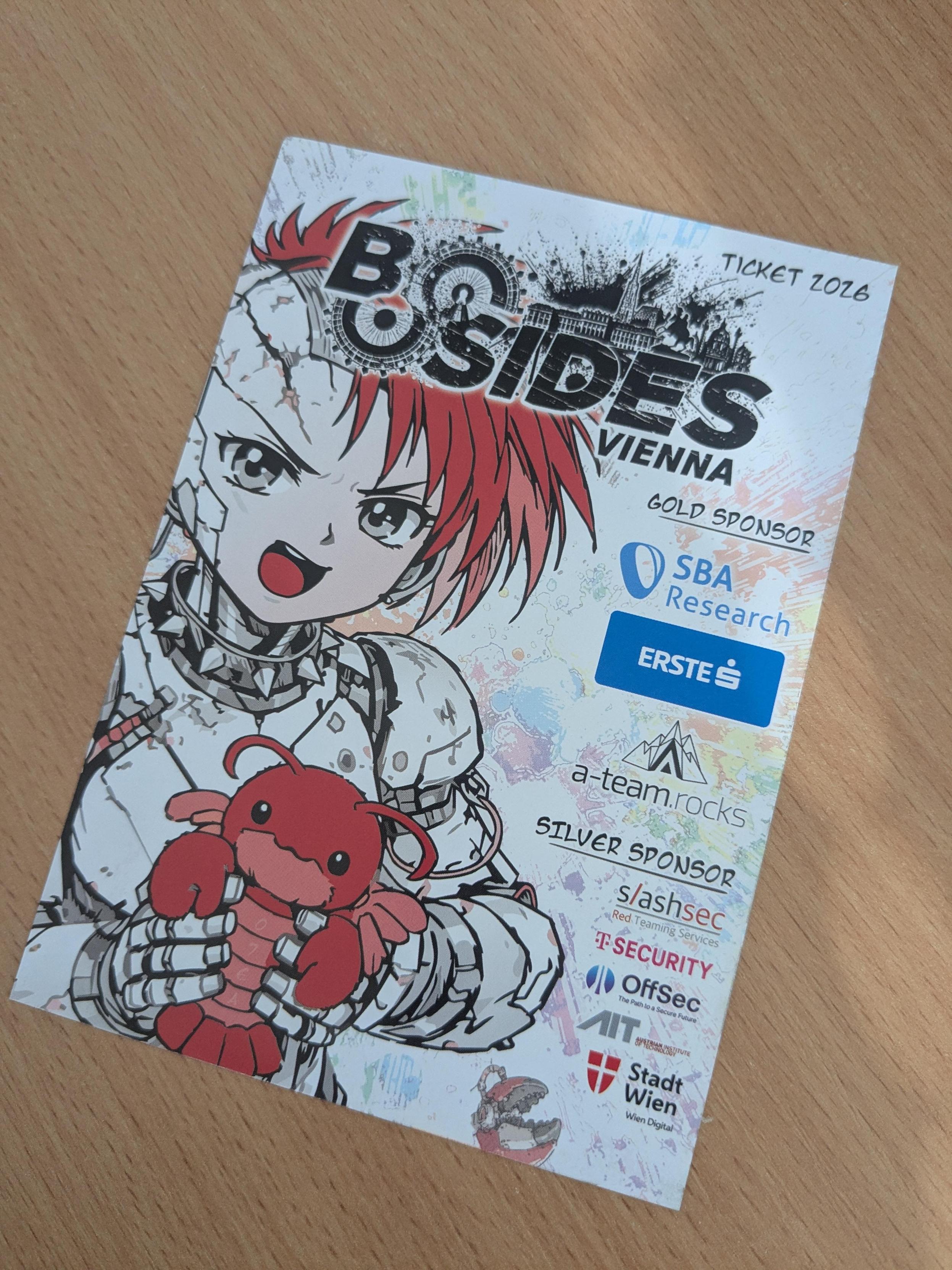 A printed conference ticket lies diagonally on a light wood table. The ticket features a colorful anime-style illustration of a red-haired cyborg character with a cracked face and mechanical arms, smiling while holding a bright red cartoon lobster plush. Across the top, large bold text reads "BSides Vienna," with "Ticket 2026" handwritten in the upper-right corner. The right side lists sponsor logos, including SBA Research and ERSTE as Gold Sponsors, followed by several Silver Sponsors. The background has a white base with colorful paint-splash accents and a black city skyline graphic behind the event title. The overall design is vibrant, playful, and cyberpunk-inspired.