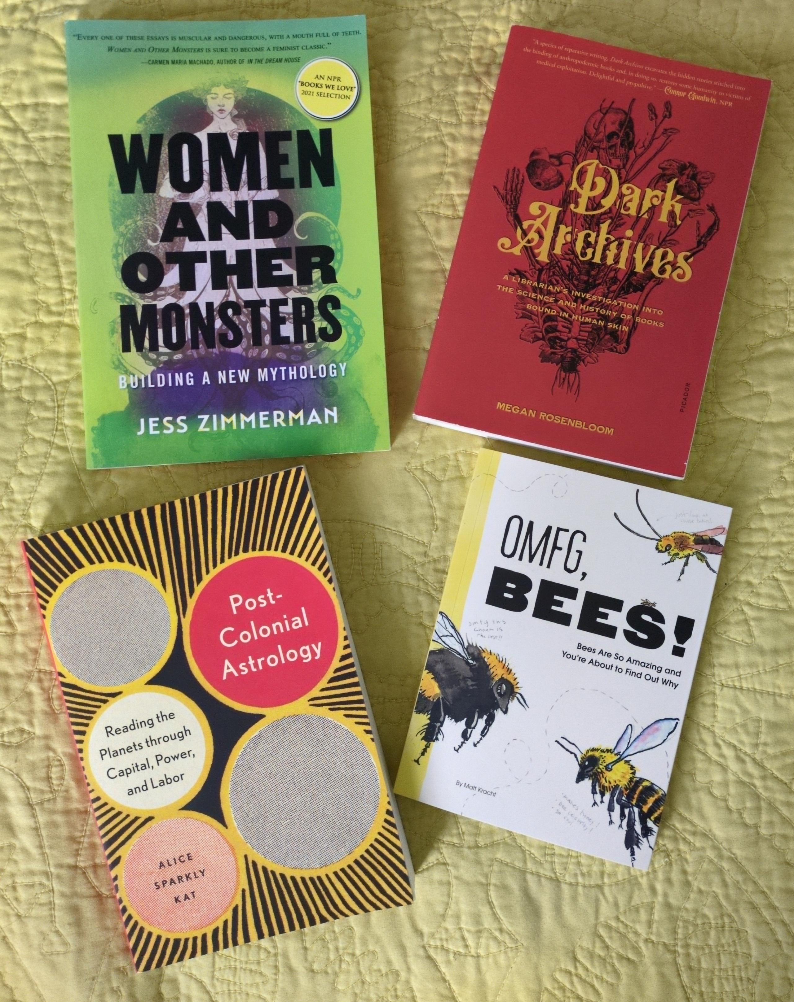 Photo of four books laid out on a yellow blanket: Jess Zimmerman's "Women and Other Monsters: Building a New Mythology", Megan Rosenbloom's "Dark Archives: A Librarian's Investigation into the Science and History of Books Bound in Human Skin", Alice Sparkly Kat's "Post-Colonial Astrology: Reading the Planets through Capital, Power, and Labor", and Matt Kracht's "OMFG, Bees! Bees Are So Amazing and You're About to Find Out Why"