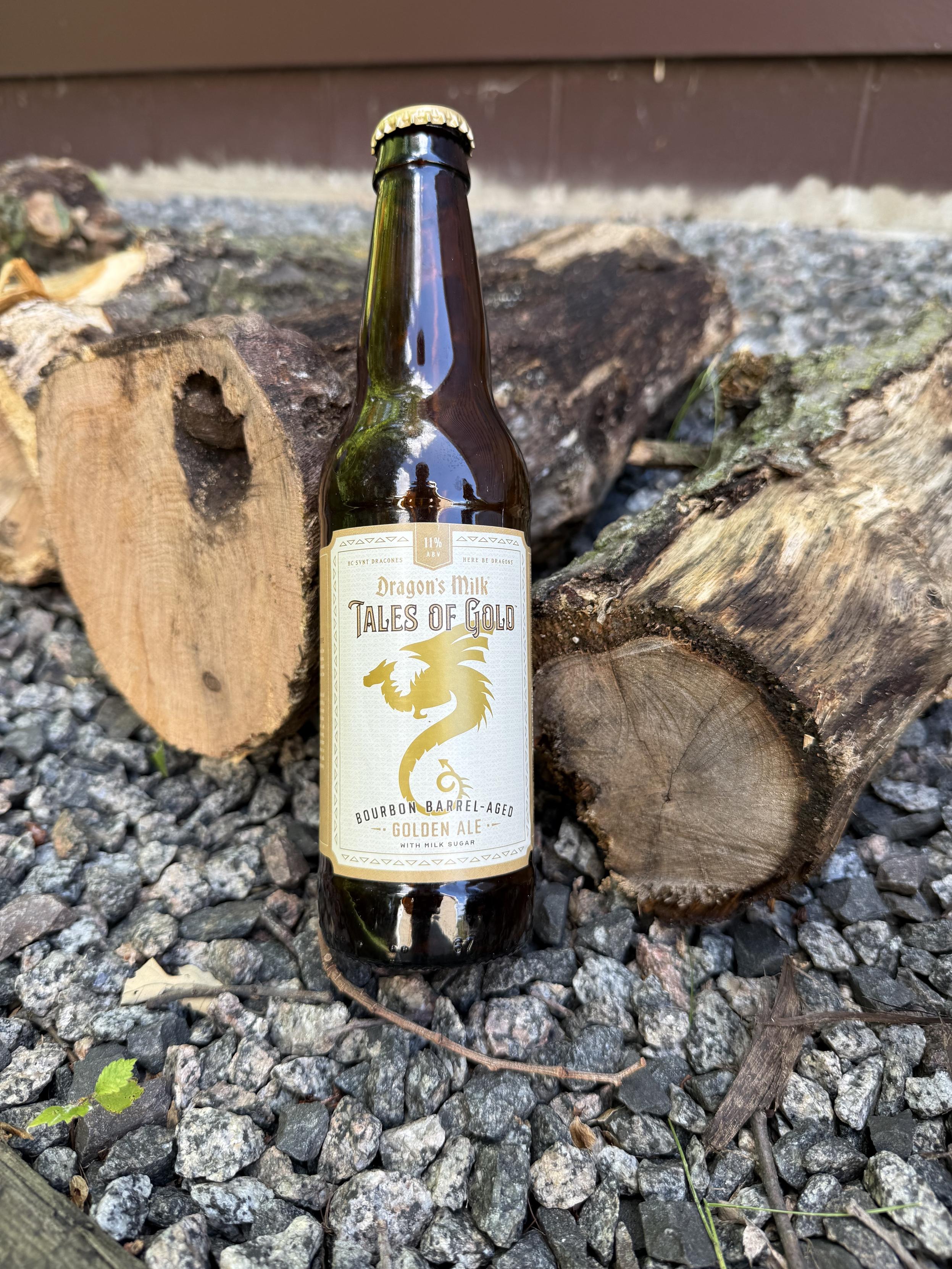A brown bottle with a white label has gold lettering and a dragon in silhouette. it is leaned against some cut oak and birch wood on a bed of crushed granite stones