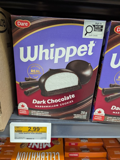 The image shows a shelf in a store with several boxes of Dare Whippet Dark Chocolate Marshmallow Cookies. The main focus is on a box in the center, which is prominently displayed. The box has a purple background with a stylized wave of dark brown and gold. The brand name "Whippet" is written in large, white, sans-serif letters across the top half of the box. Below the brand name, there's a circular badge that says "MADE WITH REAL CHOCOLATE" in white text.

The central image on the box is a clos…