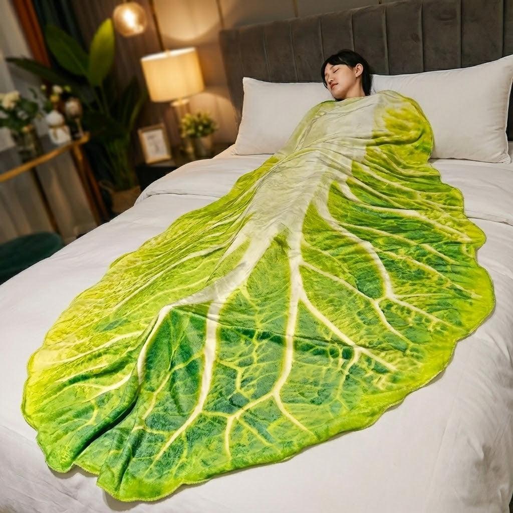Photo of someone sleeping under an actual lettuce wrap plush blanket on a bed. The throw blanket looks just like a huge leaf of lettuce.