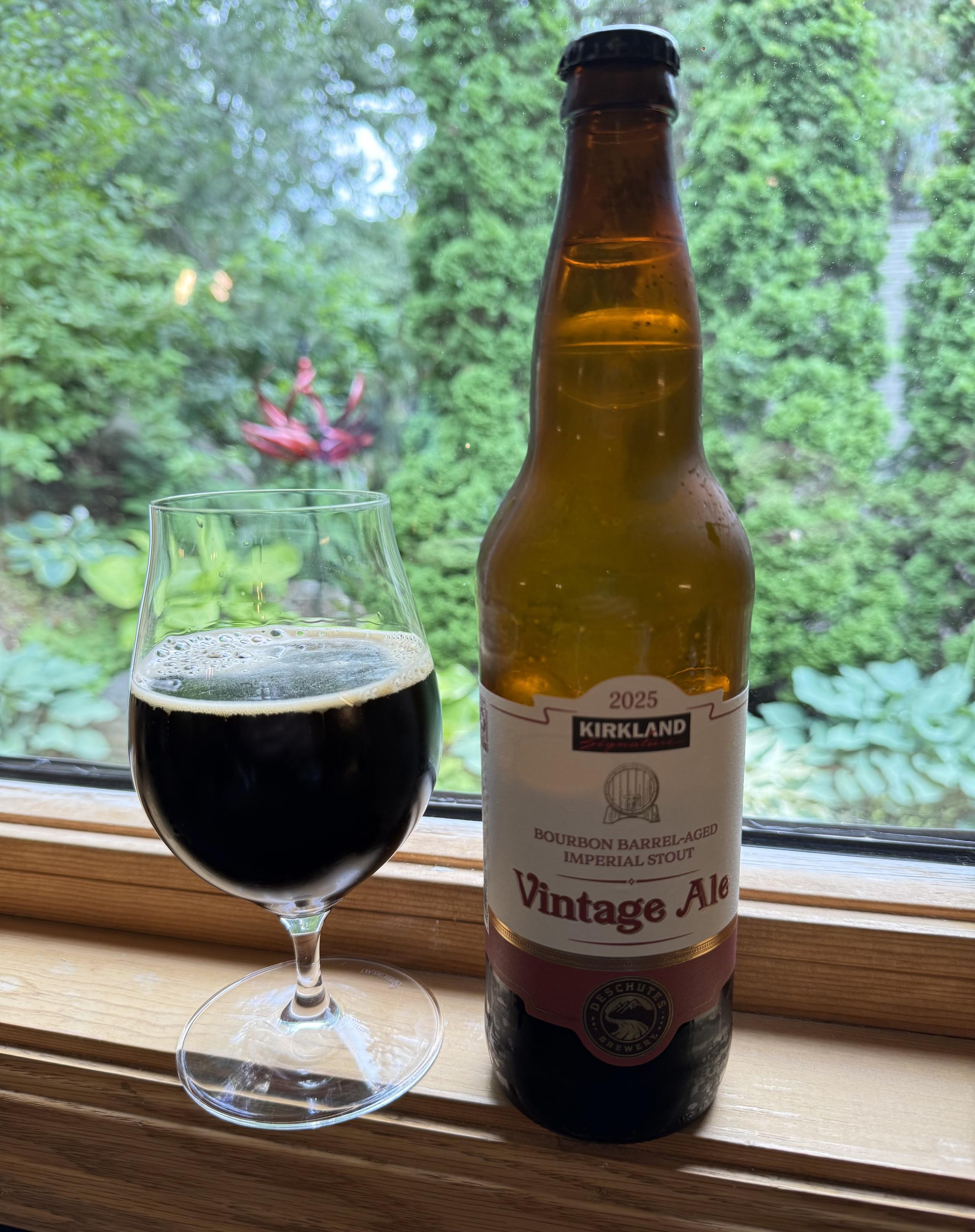 A snifter glass half full of dark beer is on a windowsill next to a tall brown bottle with a white and purple label. Tall green arborvitae are on the background