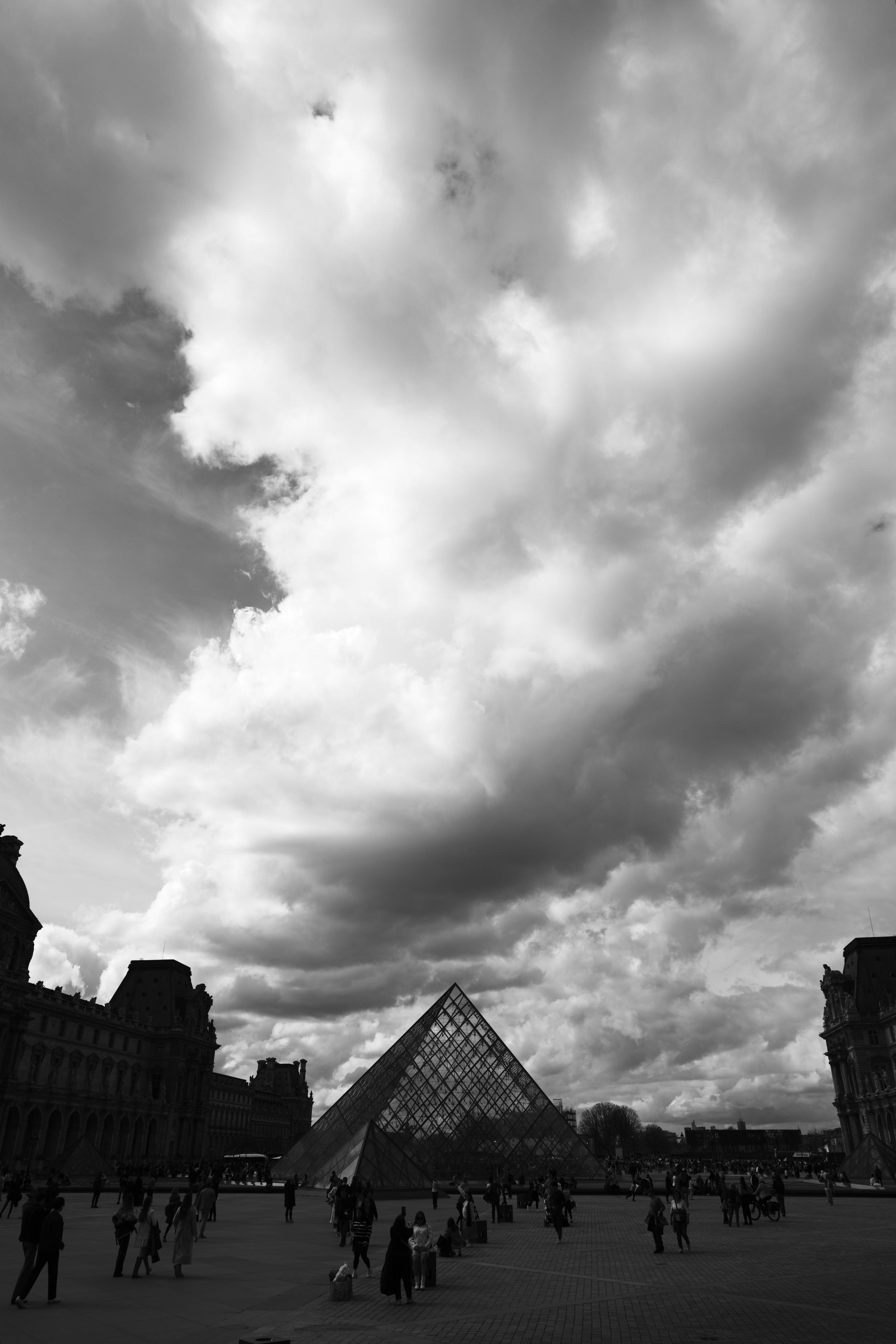 A black and white photo of dramatic clouds over the Louvre pyramid.