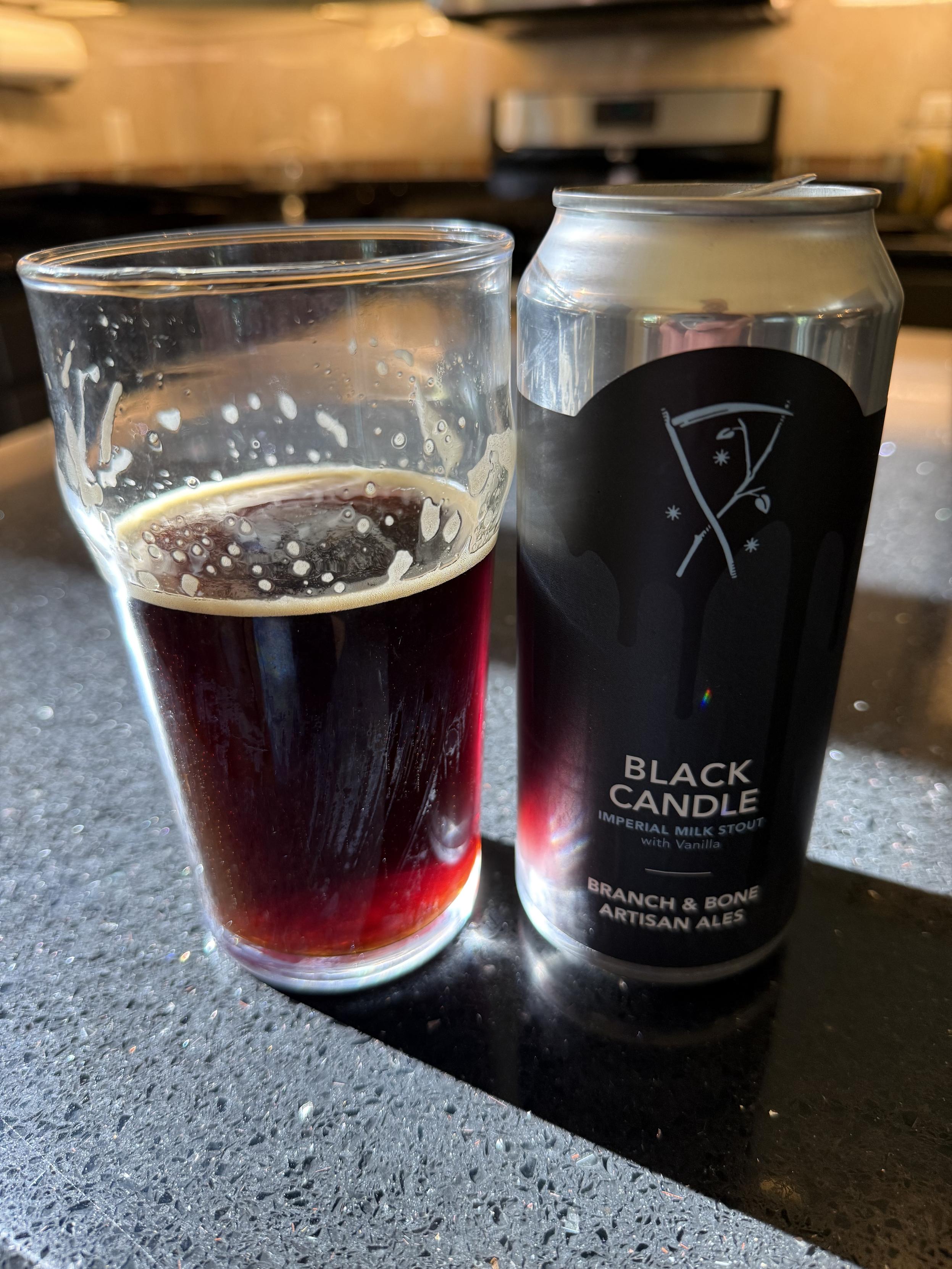 A pint glass full of almost ruby colored beer is bathed in sunlight in n a black countertop. The silver pint can has a black label