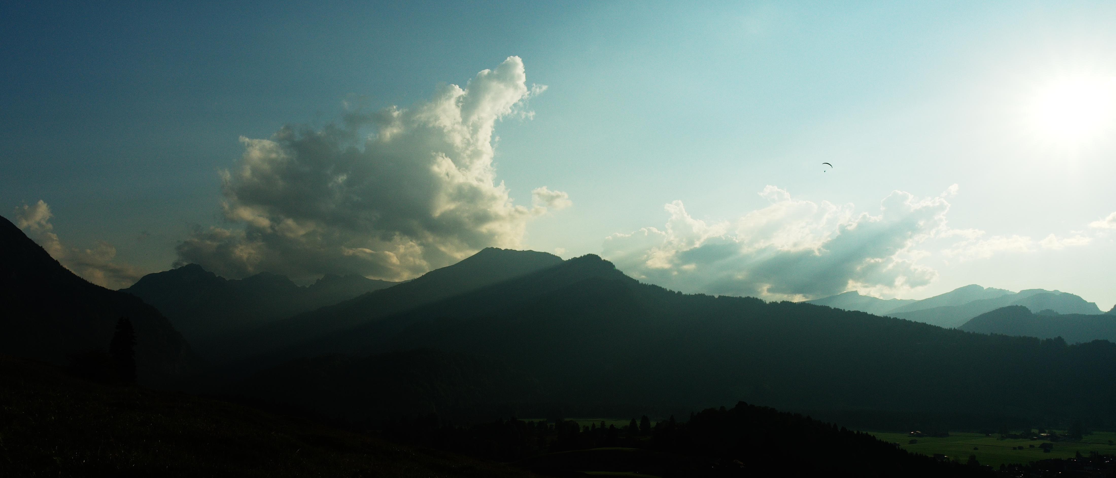 A landscape photo of mountains and clouds with sun rays scraping over some mountaintops and casting shadows. A paraglider can be seen flying in the top right quadrant.