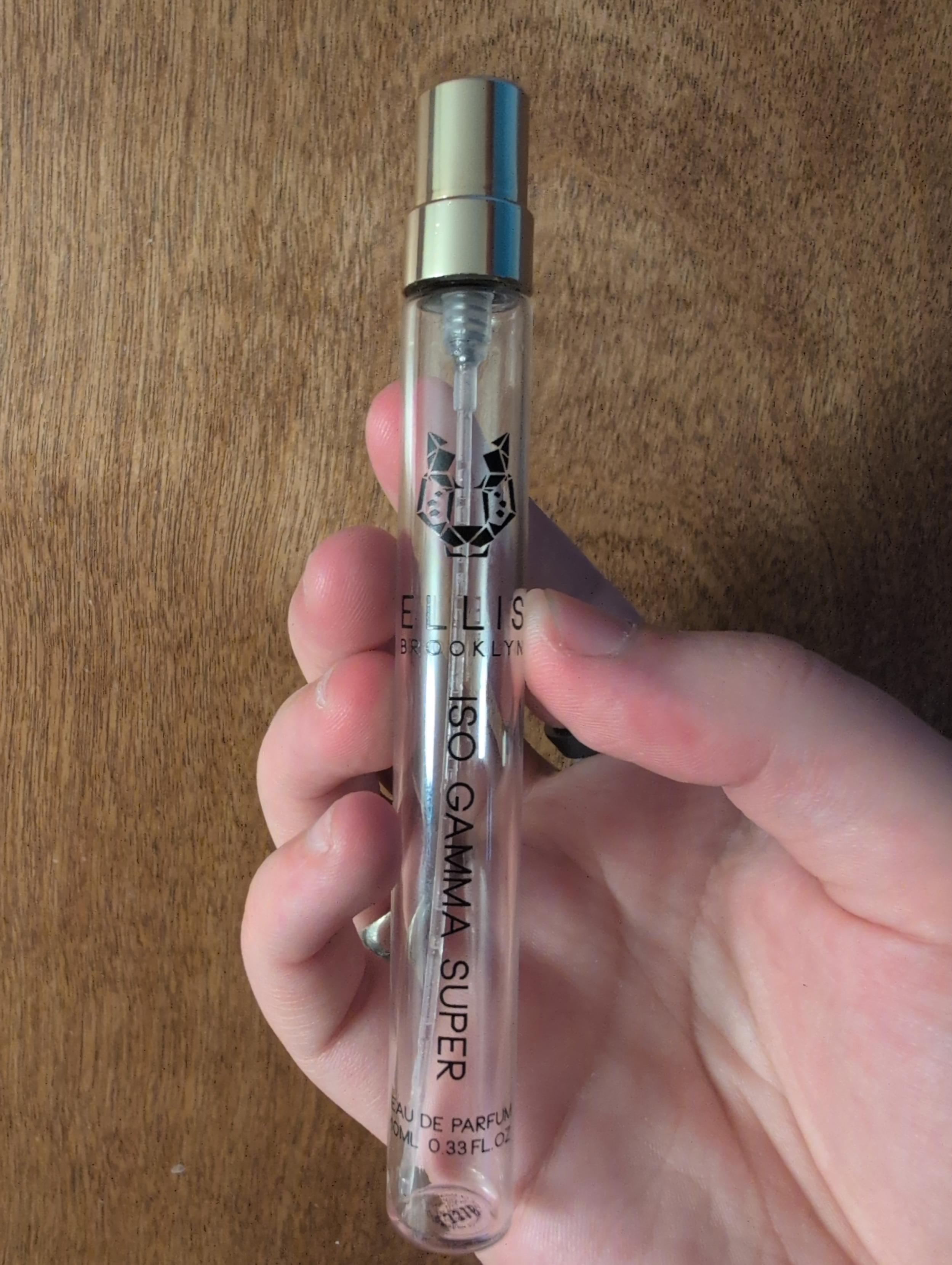 a pale slightly chubby hand holding a long thin clear glass perfume vial with a spray nozzle on top, marked "ELLIS BROOKLYN, ISO GAMMA SUPER" and a stylized tiger logo. the background is dark wood grain