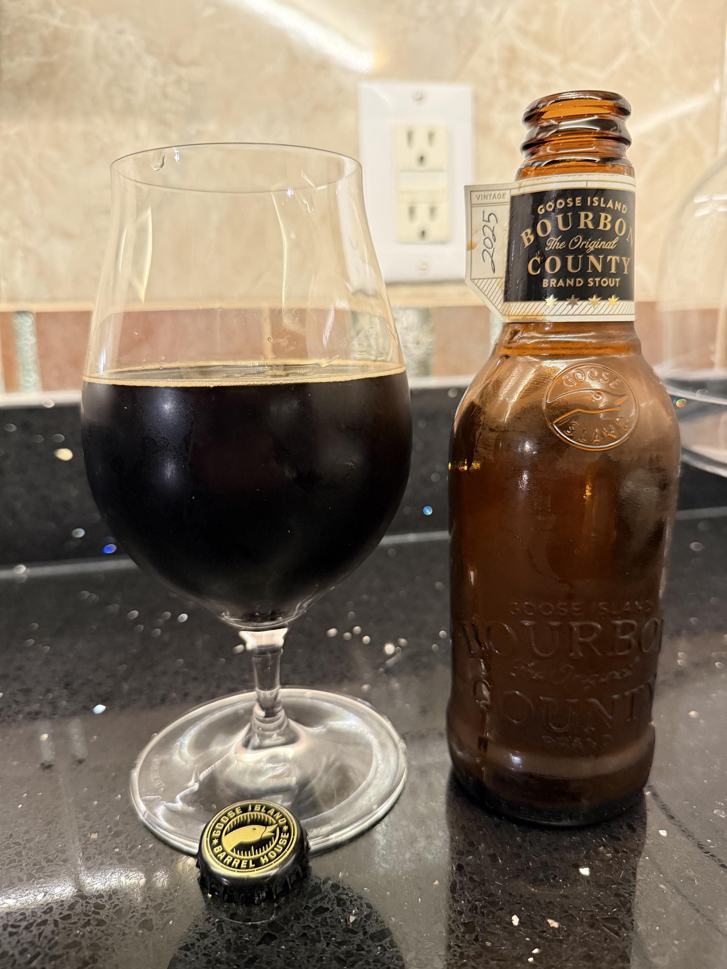 A snifter glass half full of dark beer is bathed in light, next to a short brown bottle with black and white neck label with gold lettering. A two outlet electric plug is in the background on a tiled wall