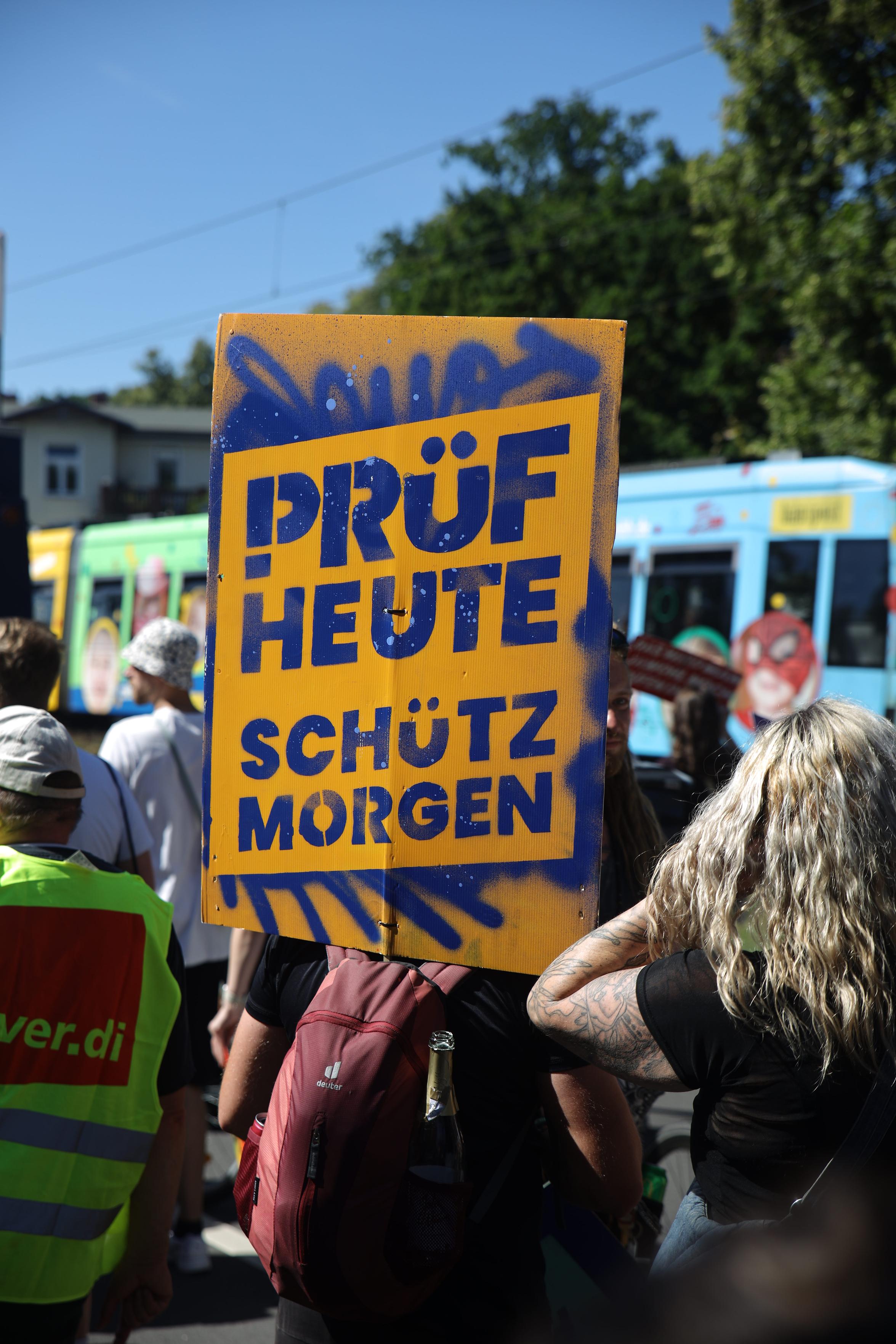 Photographie einer Demo. Ein Demoplakat liest "PRÜF HEUTE. SCHÜTZ MORGEN", dahinter eine Tram.