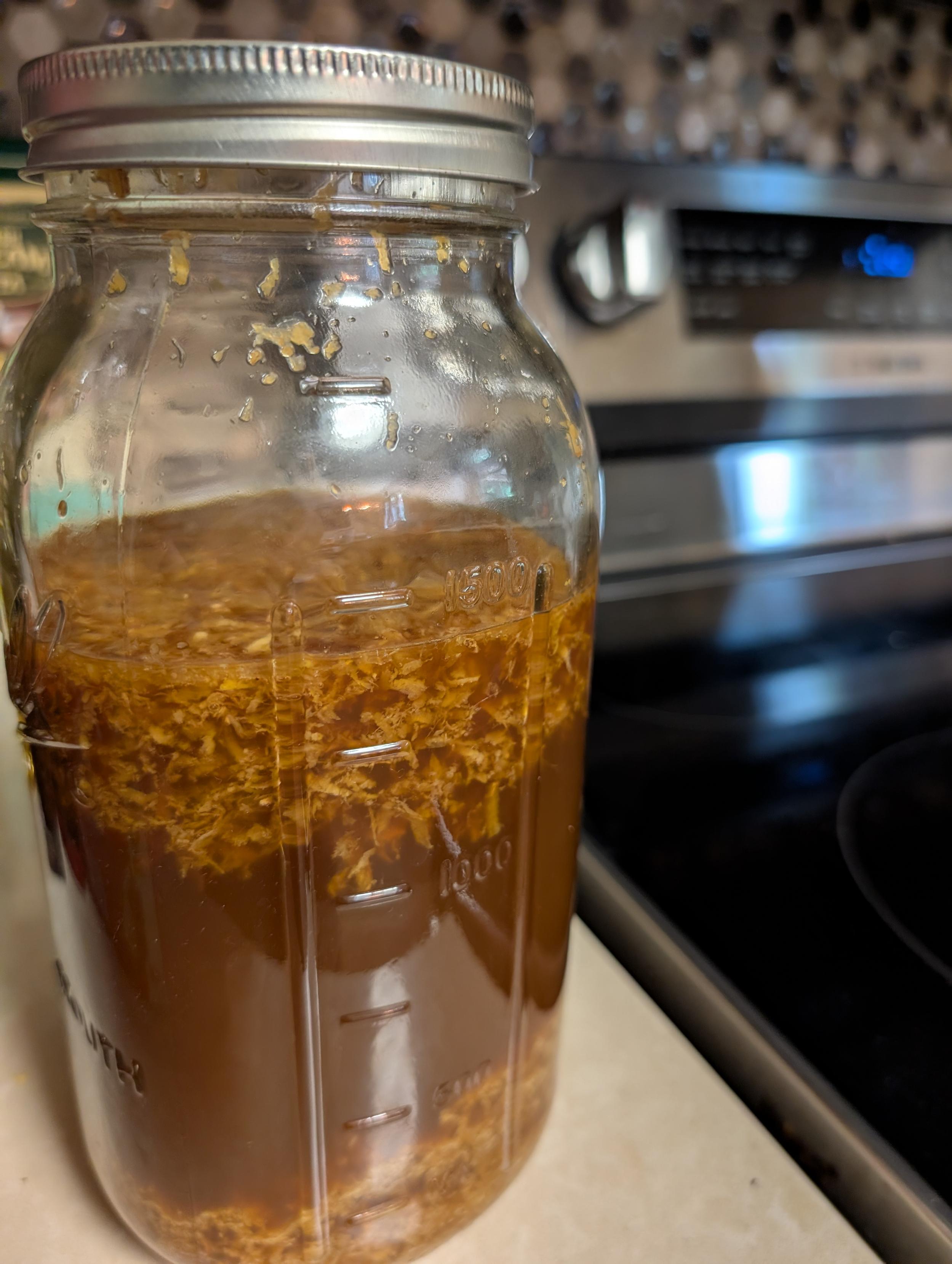 A 2qt Mason jar with a silver lid, filled with a cloudy, rich brown liquid.  Shredded ginger floats in the top quarter and rests in the bottom quarter.  In the background, a hexagonal tile kitchen backsplash and electric range.