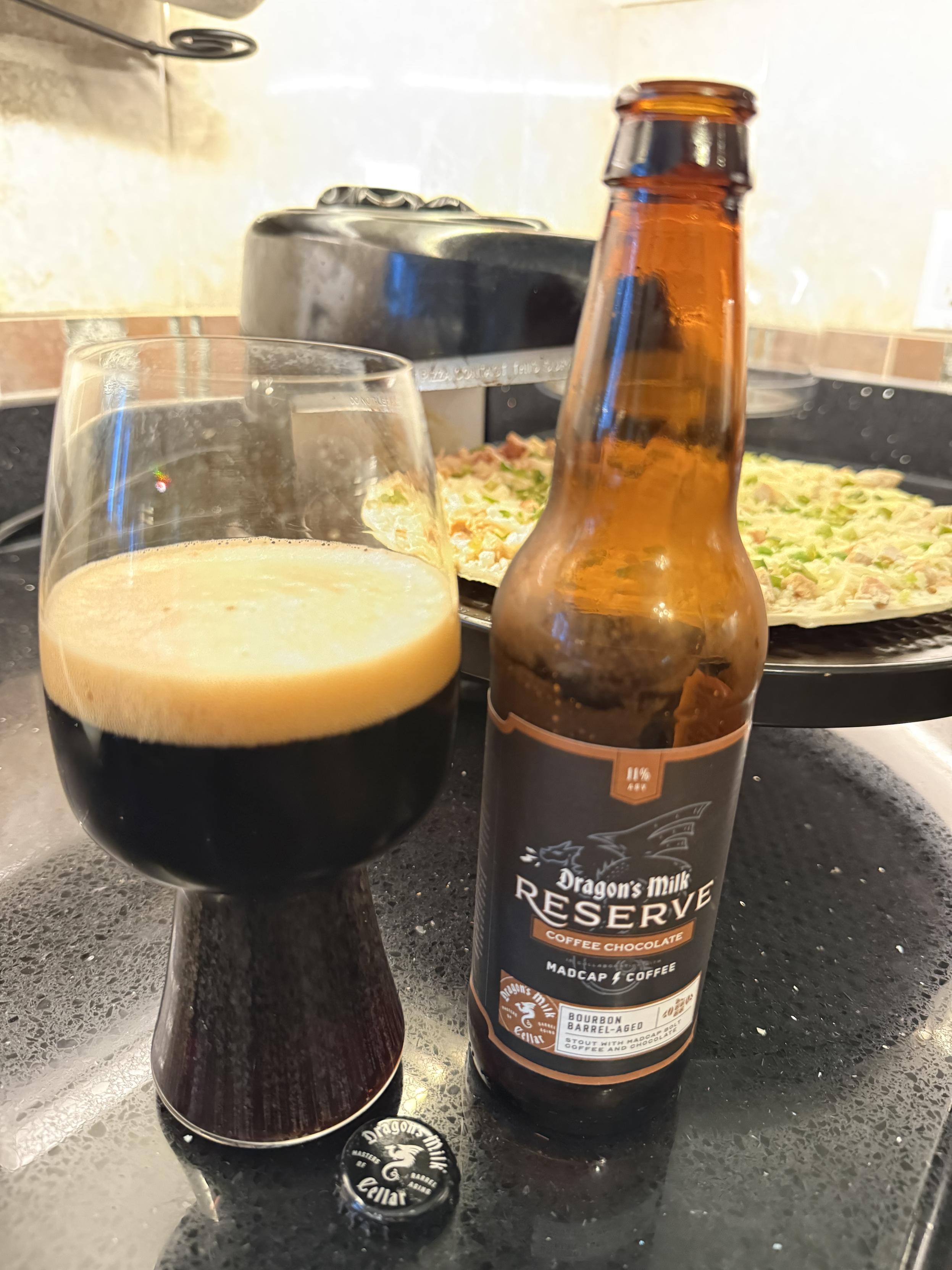 A stout glass full of dark beer with a tan head is next to a brown bottle with a black label and a white dragon in silhouette. A pizza cooks slowly in the background.