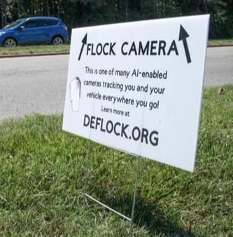 A photo of a sign stuck in grass with arrows pointing upward and text that says "Flock camera: This is one of many AI-enabled cameras tracking you and your vehicle everywhere you go! Learn more at deflock.org"