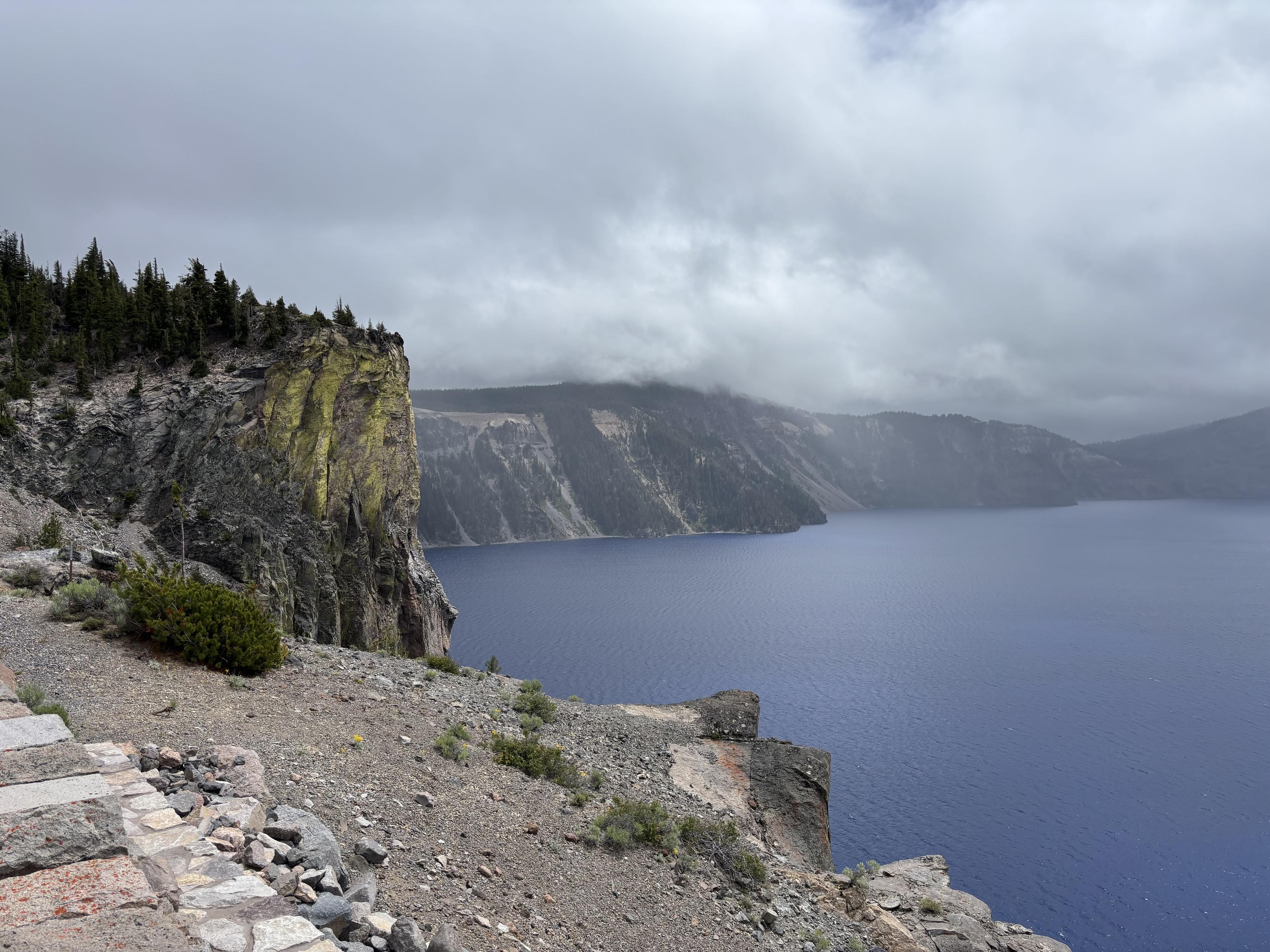 Overlooking crater lake, a section of vertical cliffs are vibrantly green yellow over the perfect blue water. Clouds roll over the rim into the lake