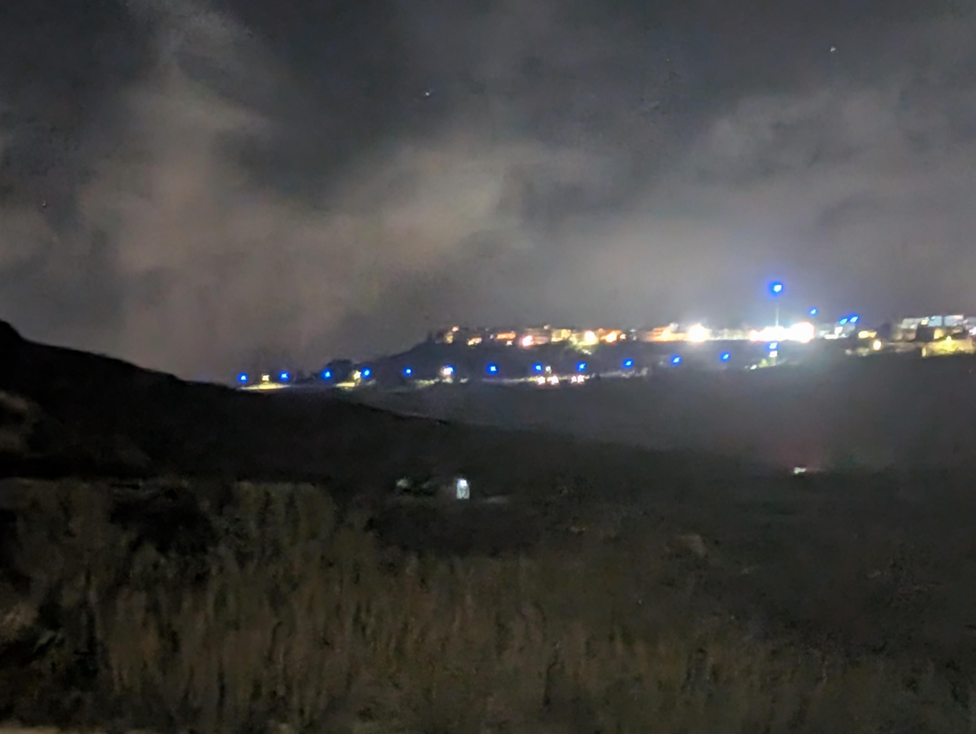 Photo of a village in the  night
Dark hills before and illuminated village and sky.
A line of blue light is visible above the vilage